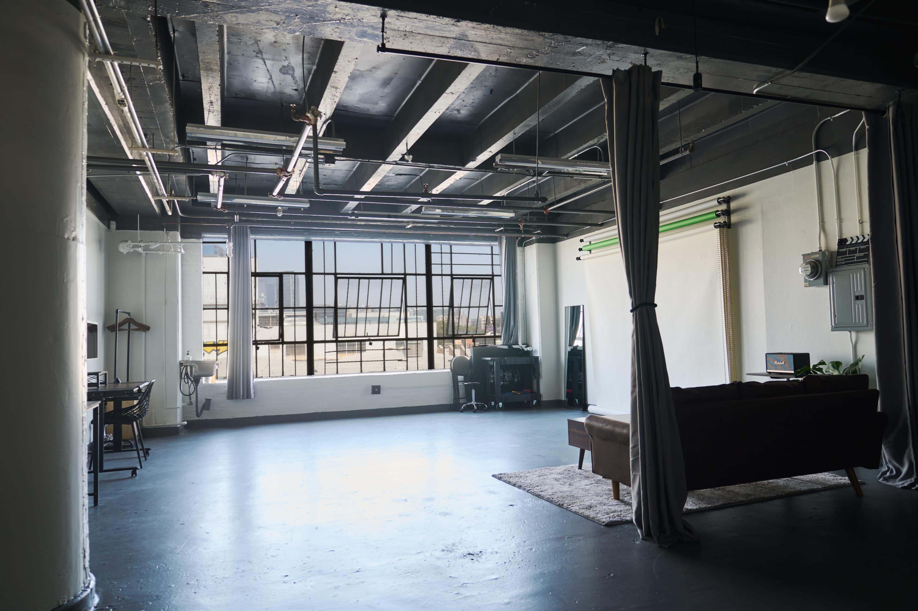 A spacious, industrial-style room with large windows, featuring a couch, a desk setup, and a projector screen against one wall.