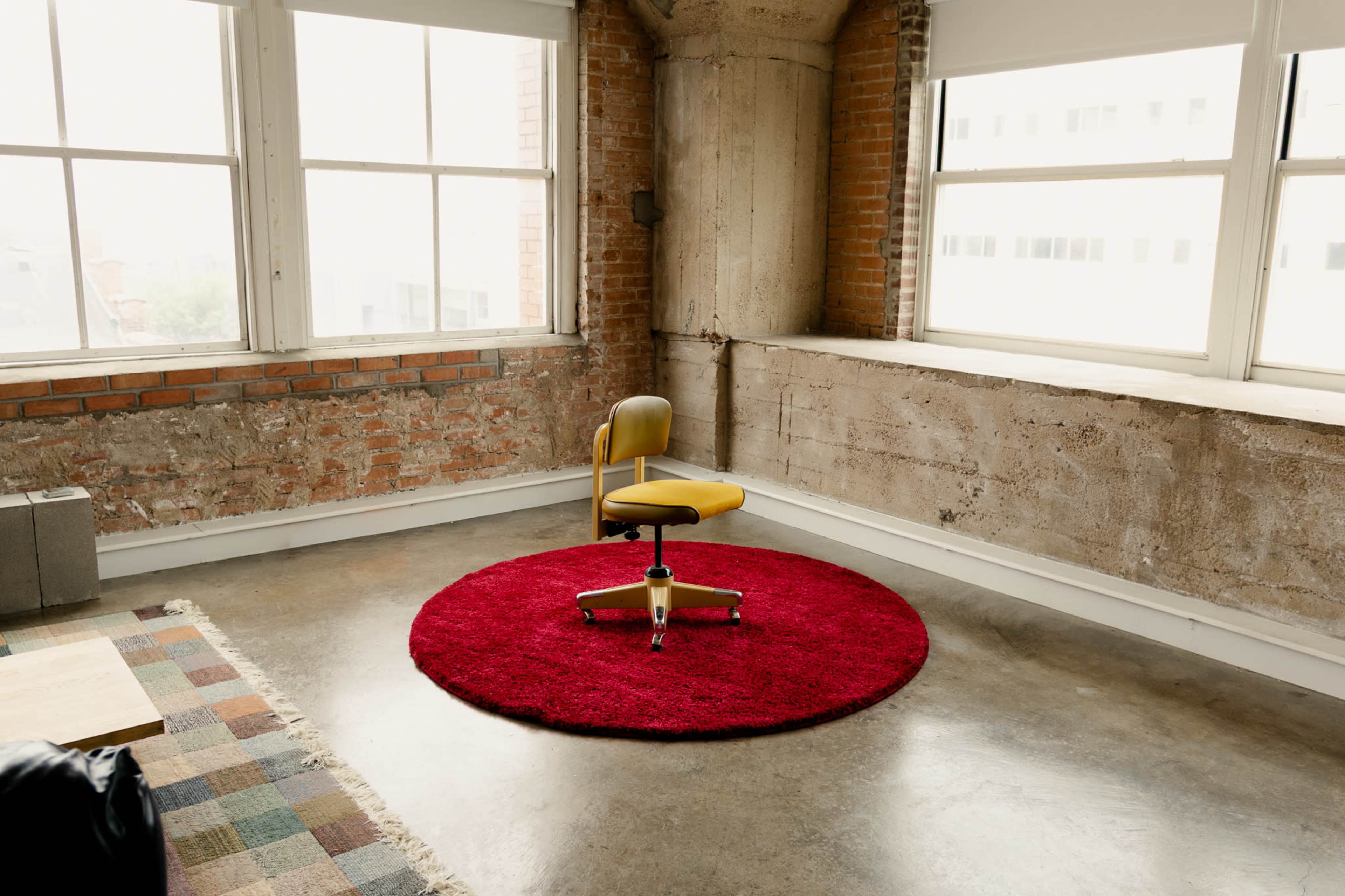 A yellow office chair sits on a red circular rug in a sunlit room with large windows and textured concrete walls.