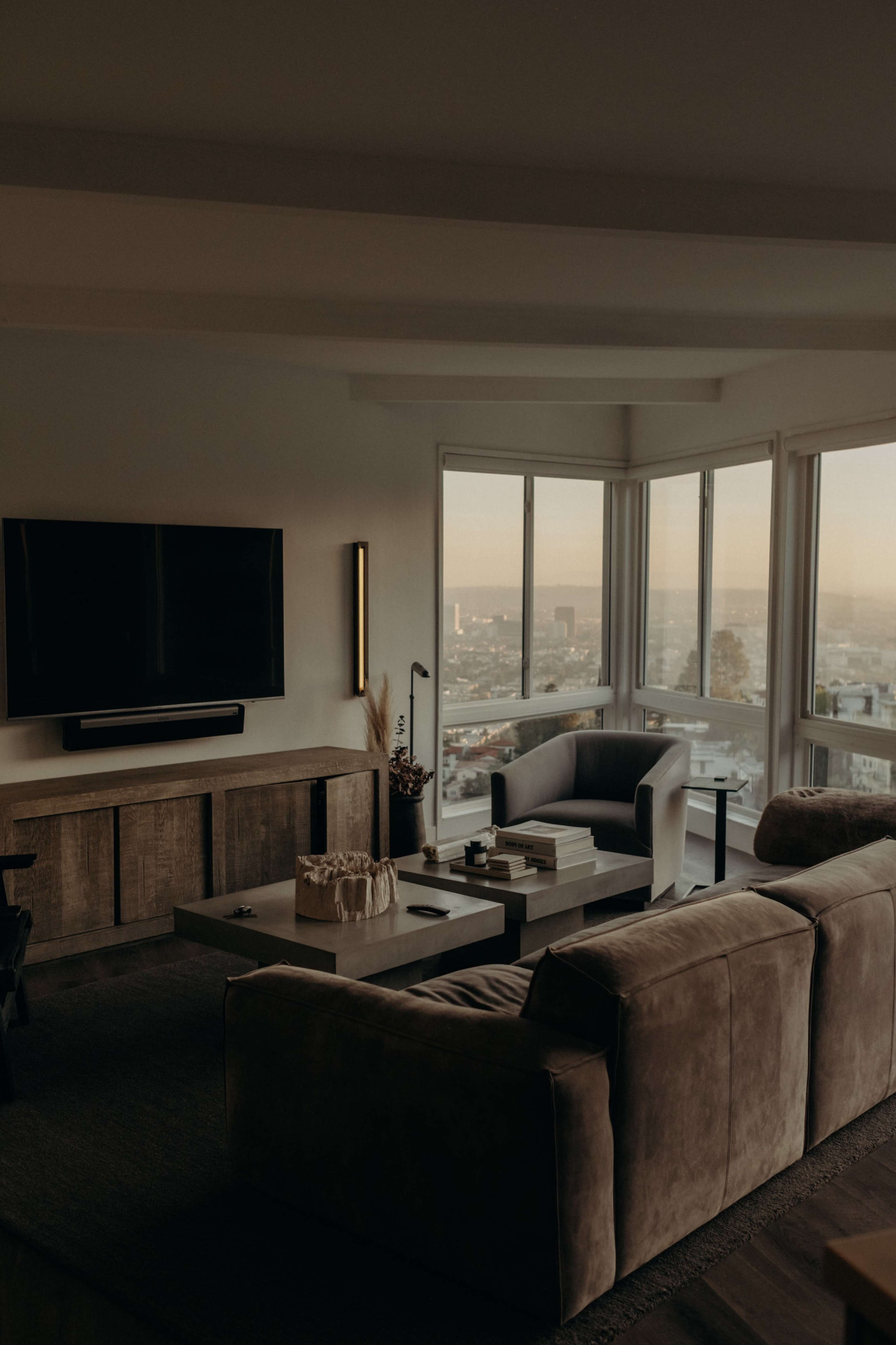A modern living room features a large window with a view of the city, a sectional sofa, and a mounted TV on a sleek cabinet.