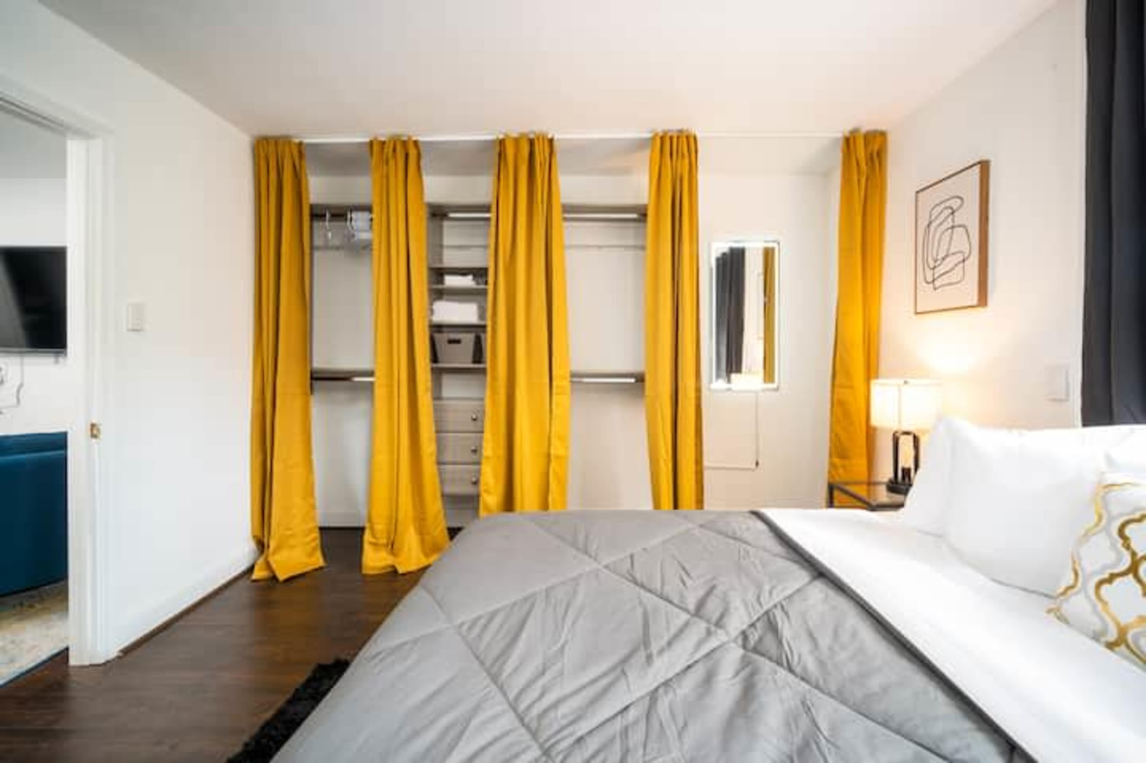 A modern bedroom features a bed with a gray quilt, yellow curtains separating a closet space, and a small lamp on a nightstand.