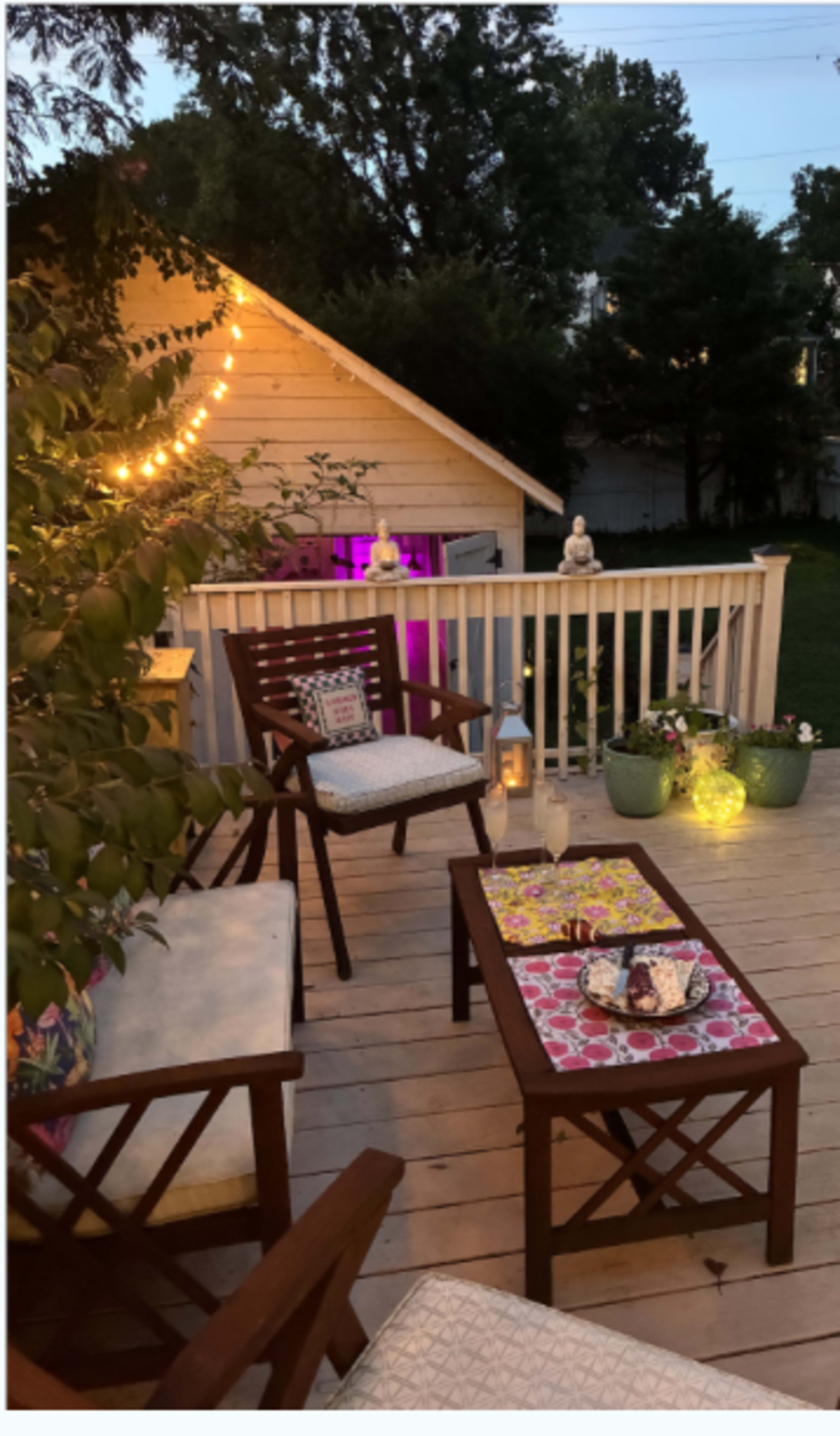 The image features a cozy deck with wooden furniture, a small table adorned with colorful decor, and decorative lights illuminating the space.
