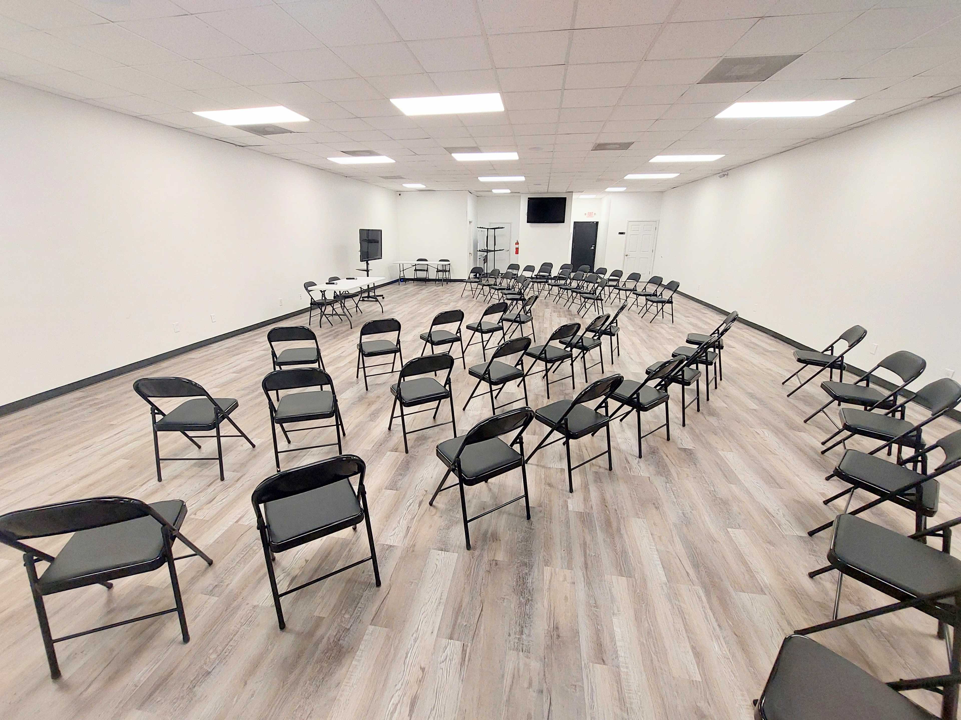 A large, empty room set up with chairs arranged in a circle, with tables and audio equipment in the background.