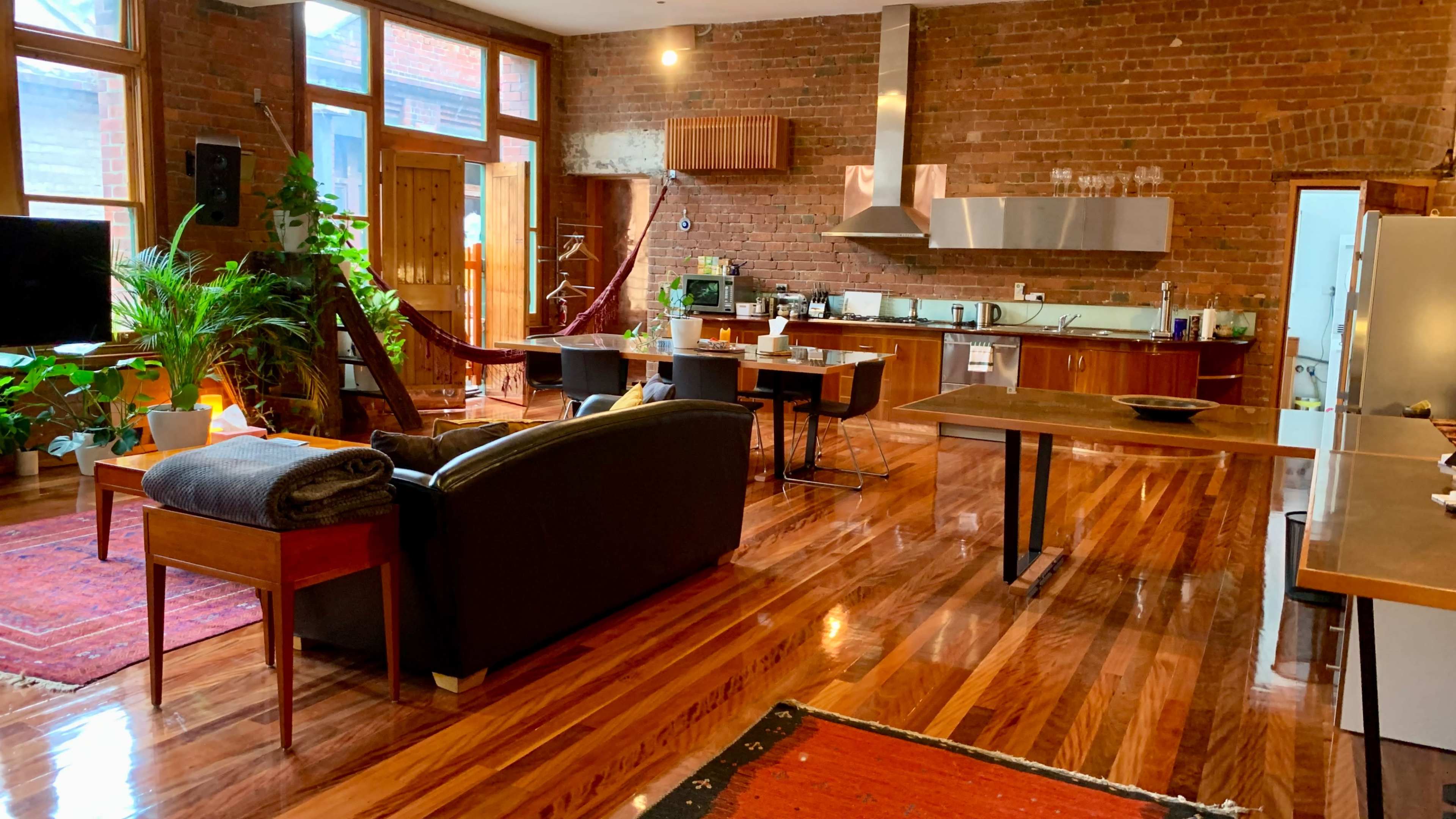 A spacious open-concept living area with brick walls, hardwood floors, a kitchen, dining area, and seating arrangements, illuminated by natural light from large windows.