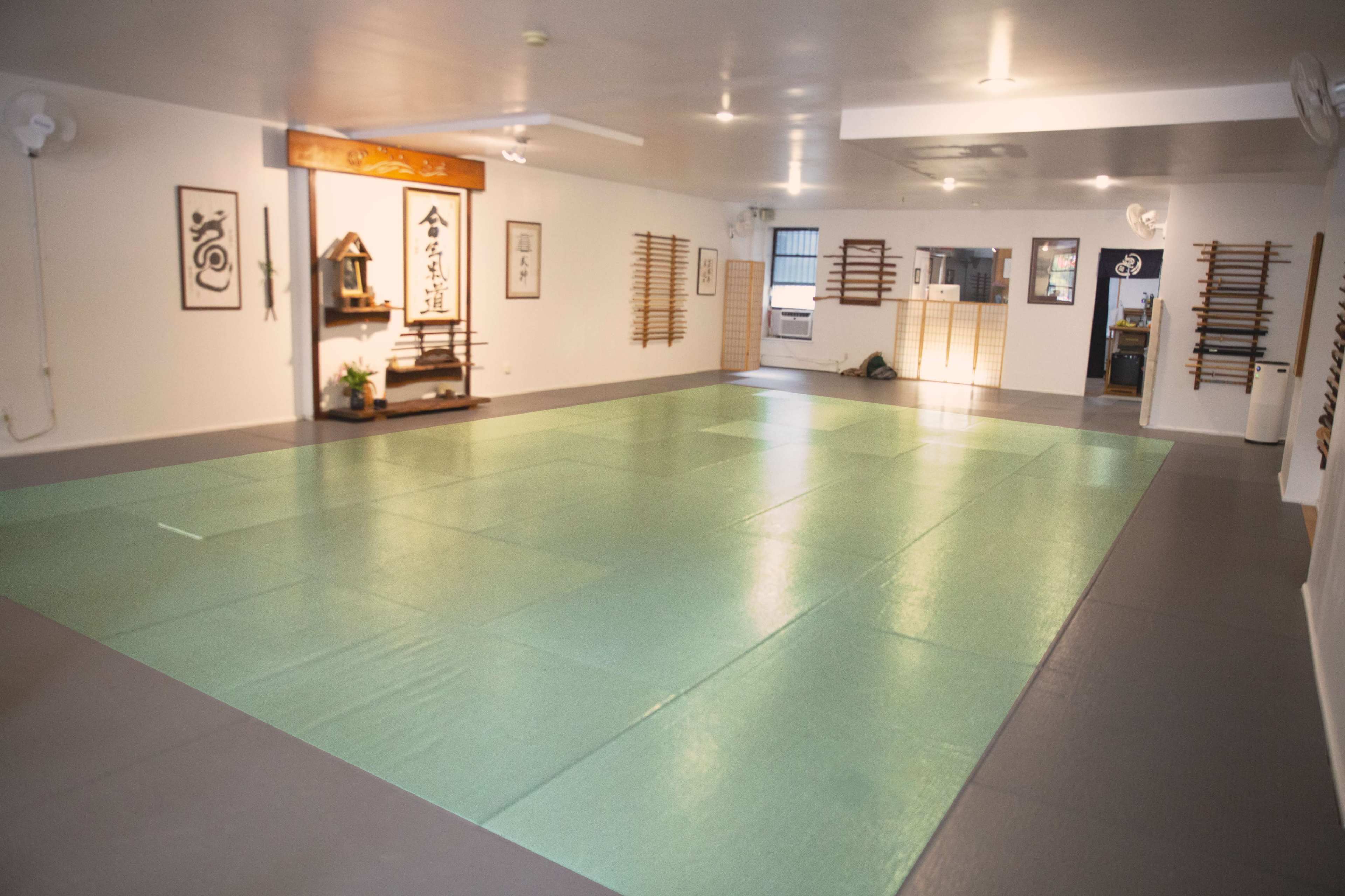 A spacious martial arts studio with green tatami mats and wooden equipment stored on walls.