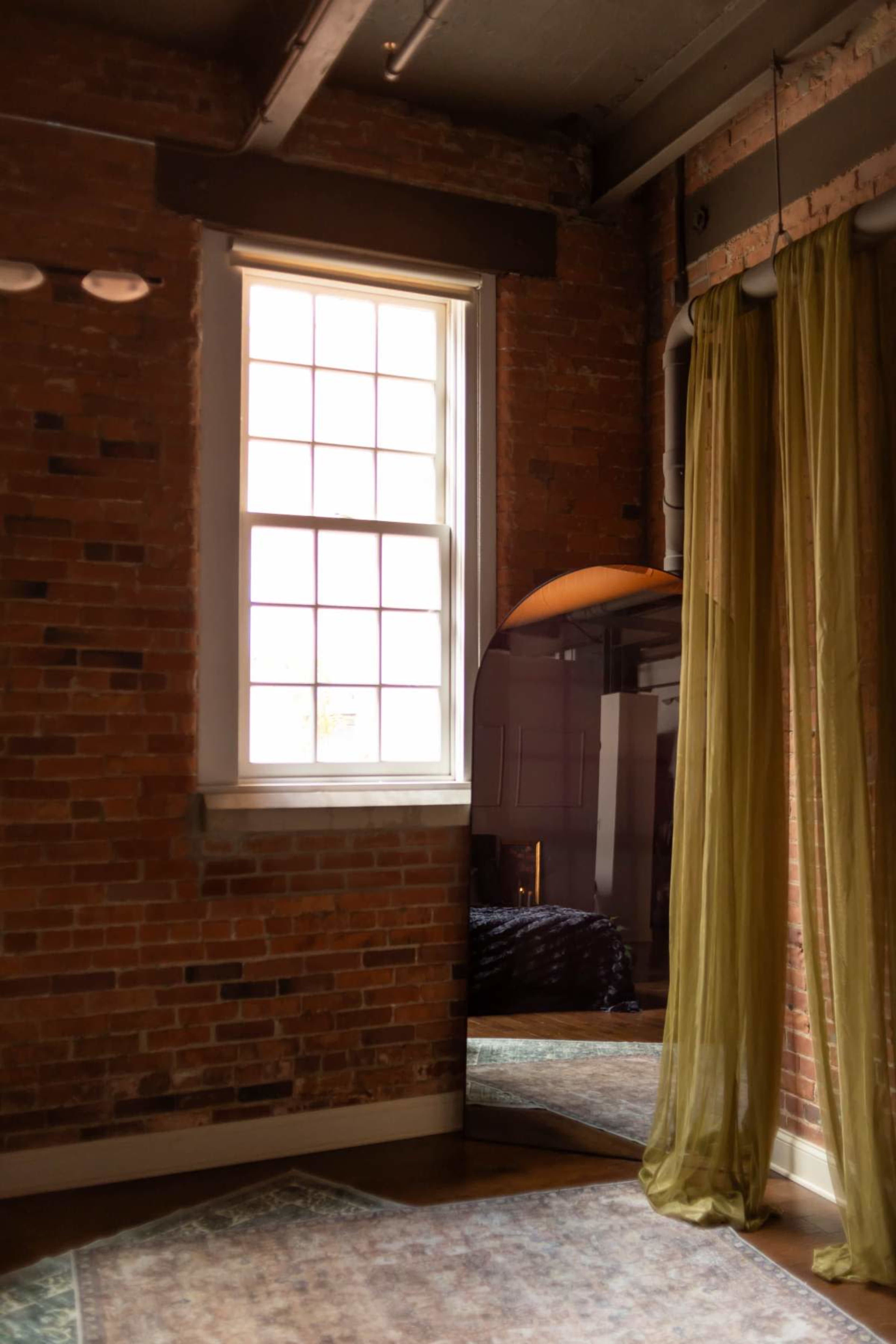 The image shows a corner of a room with exposed brick walls, a large window, a floor mirror, and sheer green curtains.