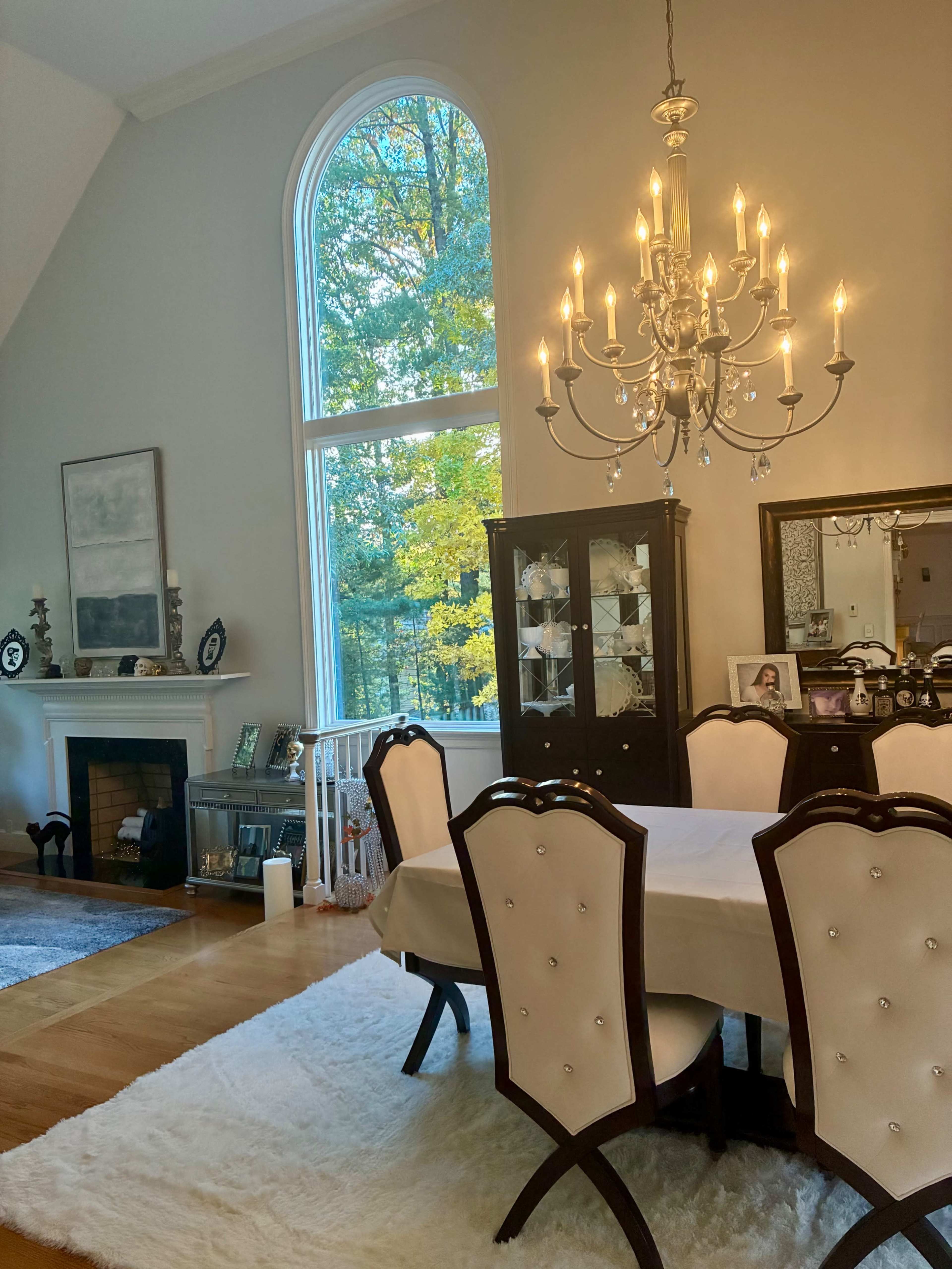 A dining area features a white table set with chairs, a chandelier above, and large windows displaying a view of trees outside.