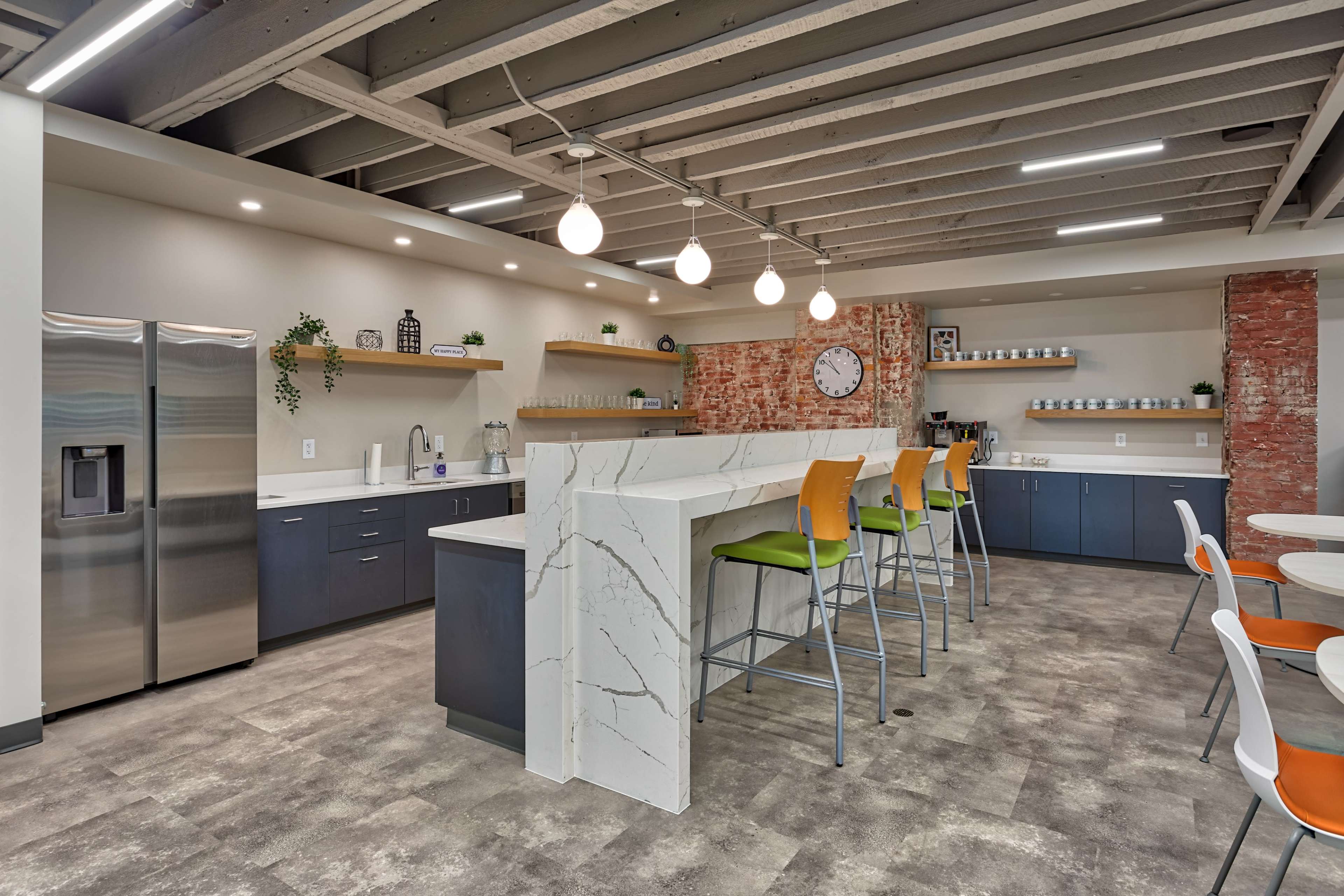 The image shows a modern kitchen area with a long bar, high stools, and a combination of open shelving and cabinetry against a brick wall.