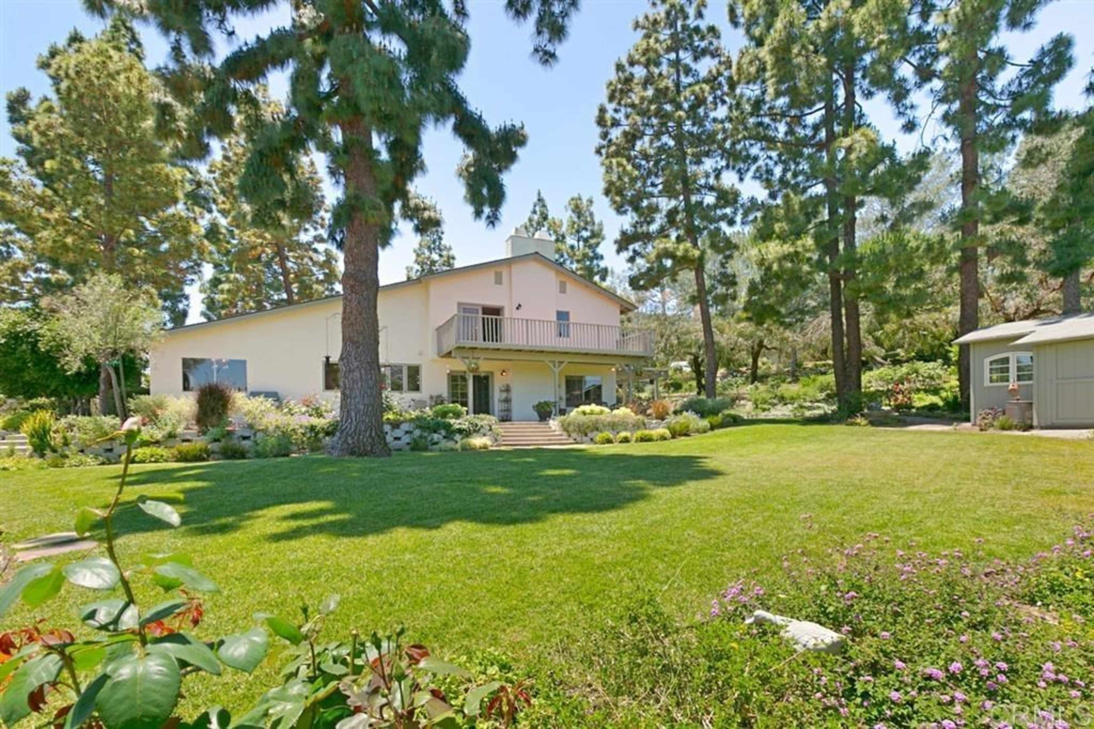 A two-story house surrounded by tall trees and a well-maintained garden with lush greenery and colorful flowers.