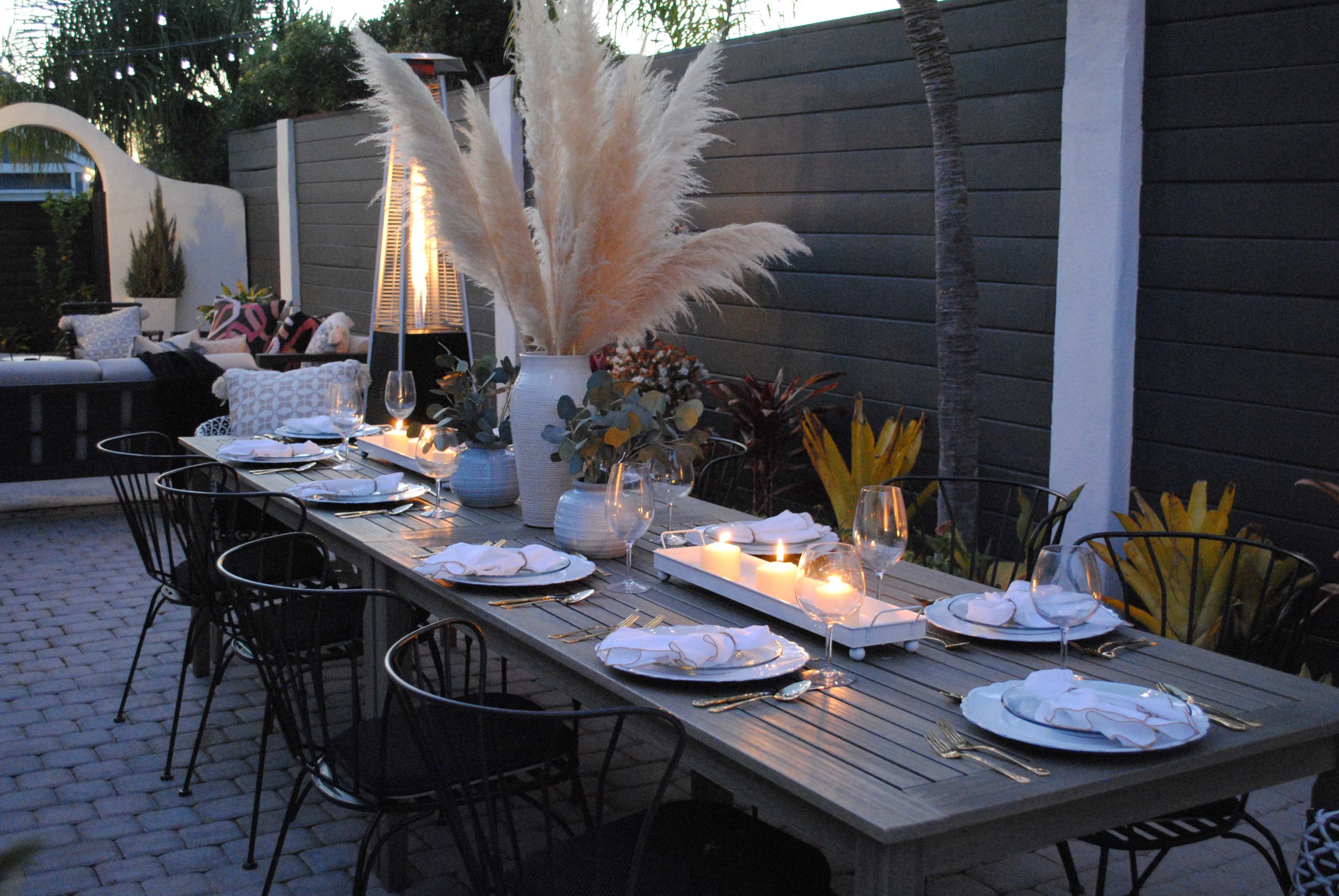 A long wooden dining table is set with white plates and glasses, flanked by black chairs, in an outdoor area adorned with decorative plants and lit by candles.
