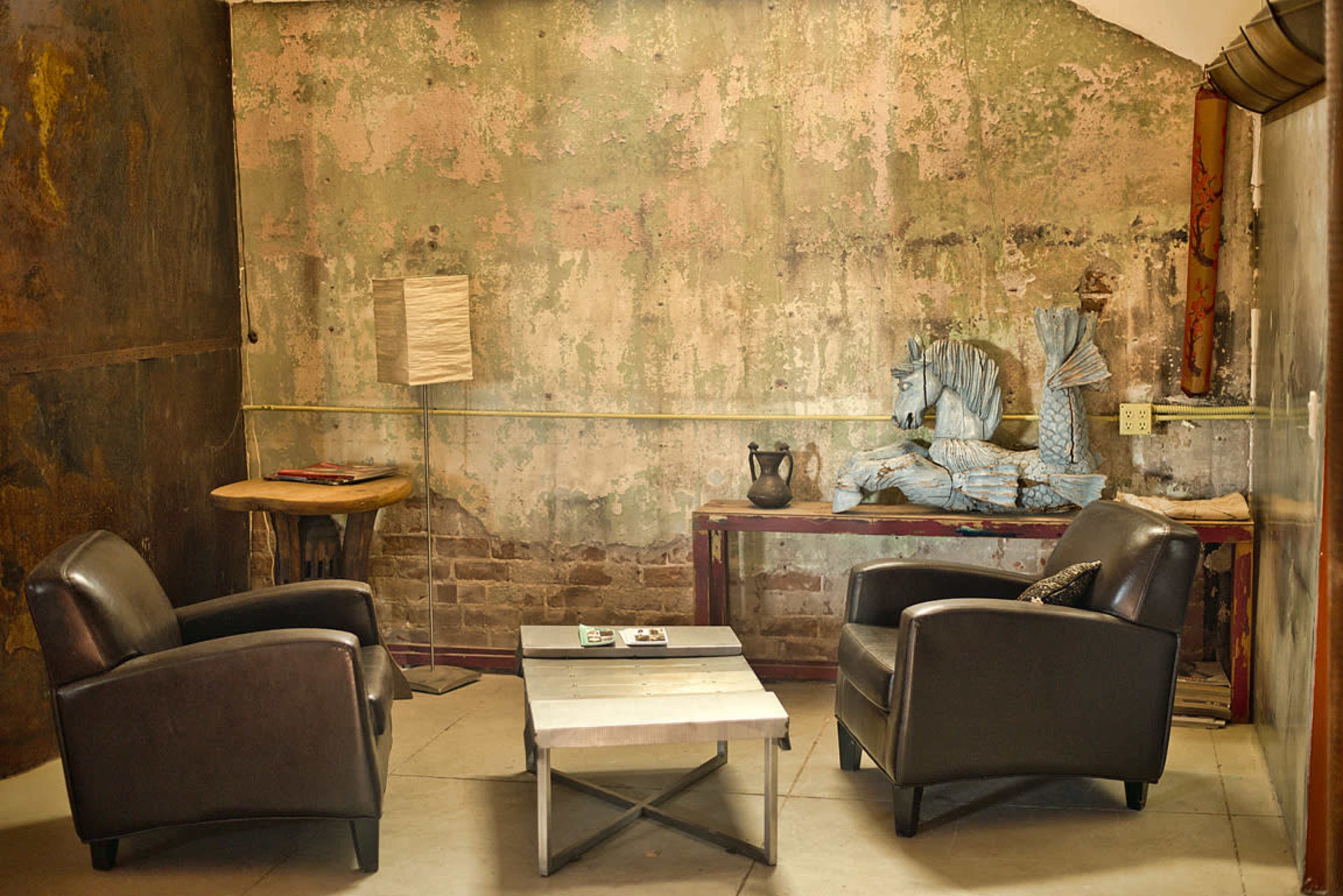 A corner of a room with peeling walls, two black armchairs facing a metal coffee table, a wooden side table with a lamp, and a decorative horse sculpture on a console.