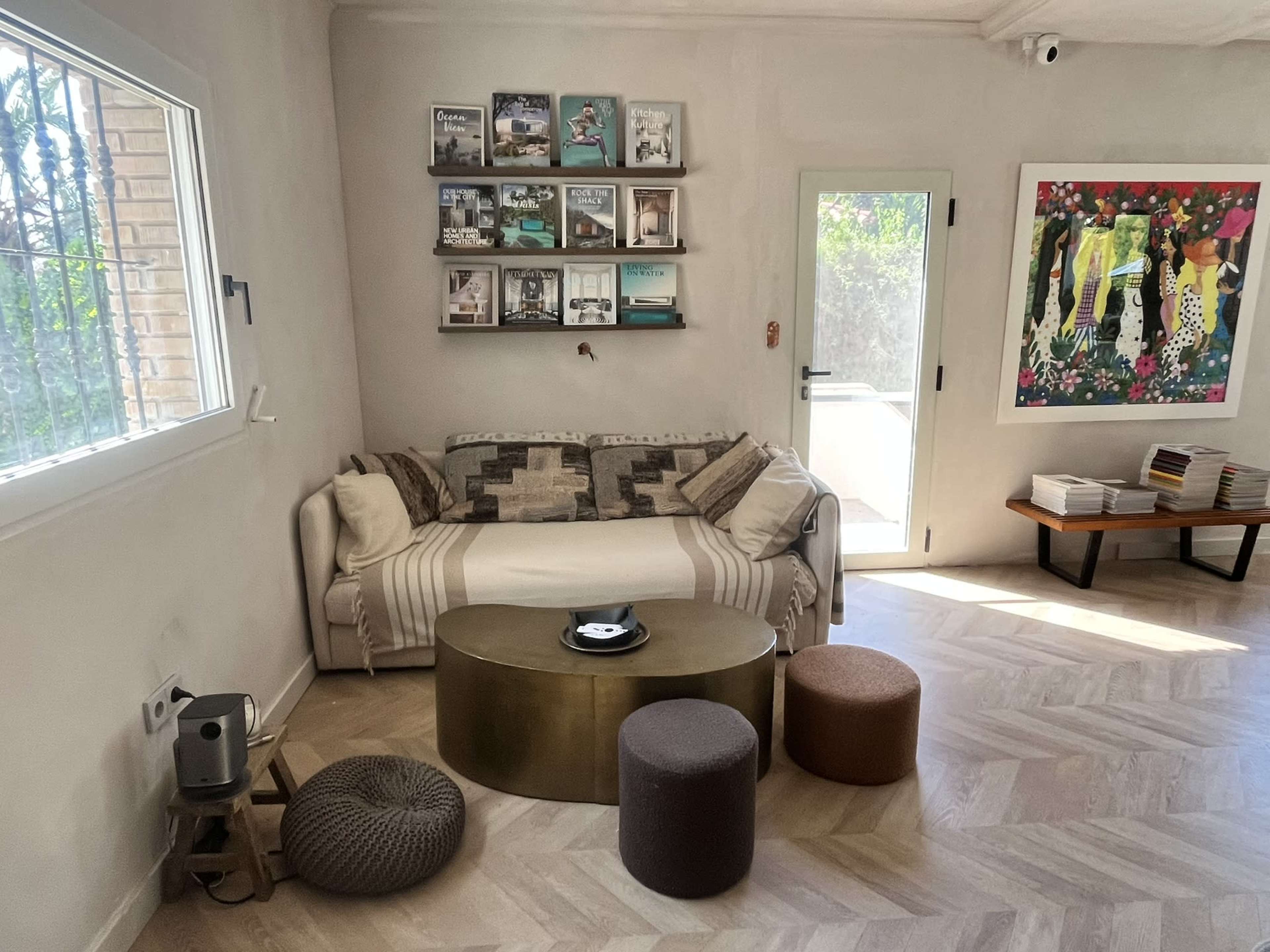 The image shows a cozy living room with a light-colored couch, a round coffee table, colorful ottomans, and a gallery wall with framed photos and artwork.