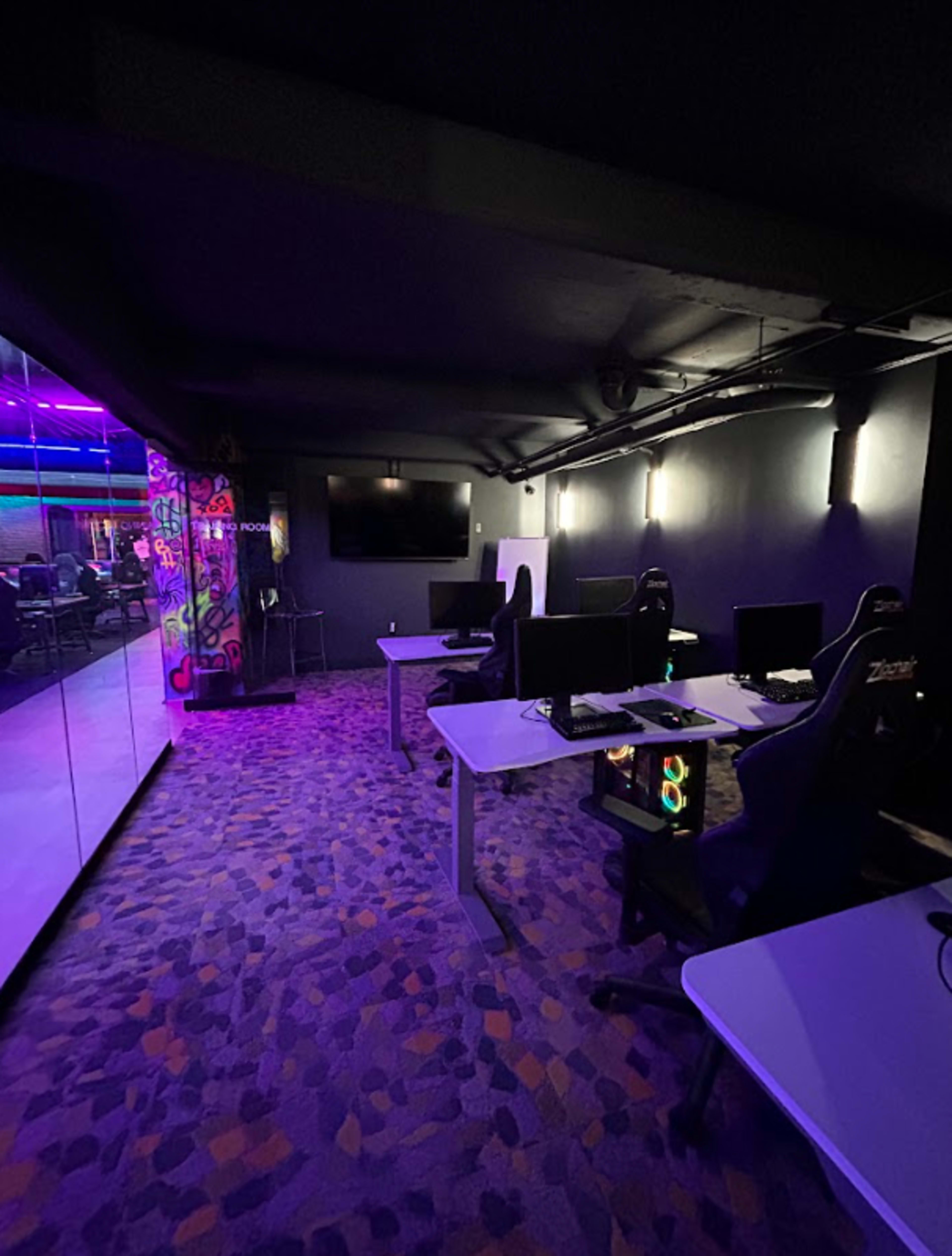 The image shows a dimly lit gaming room with several computer desks, gaming chairs, and a large screen on the wall.