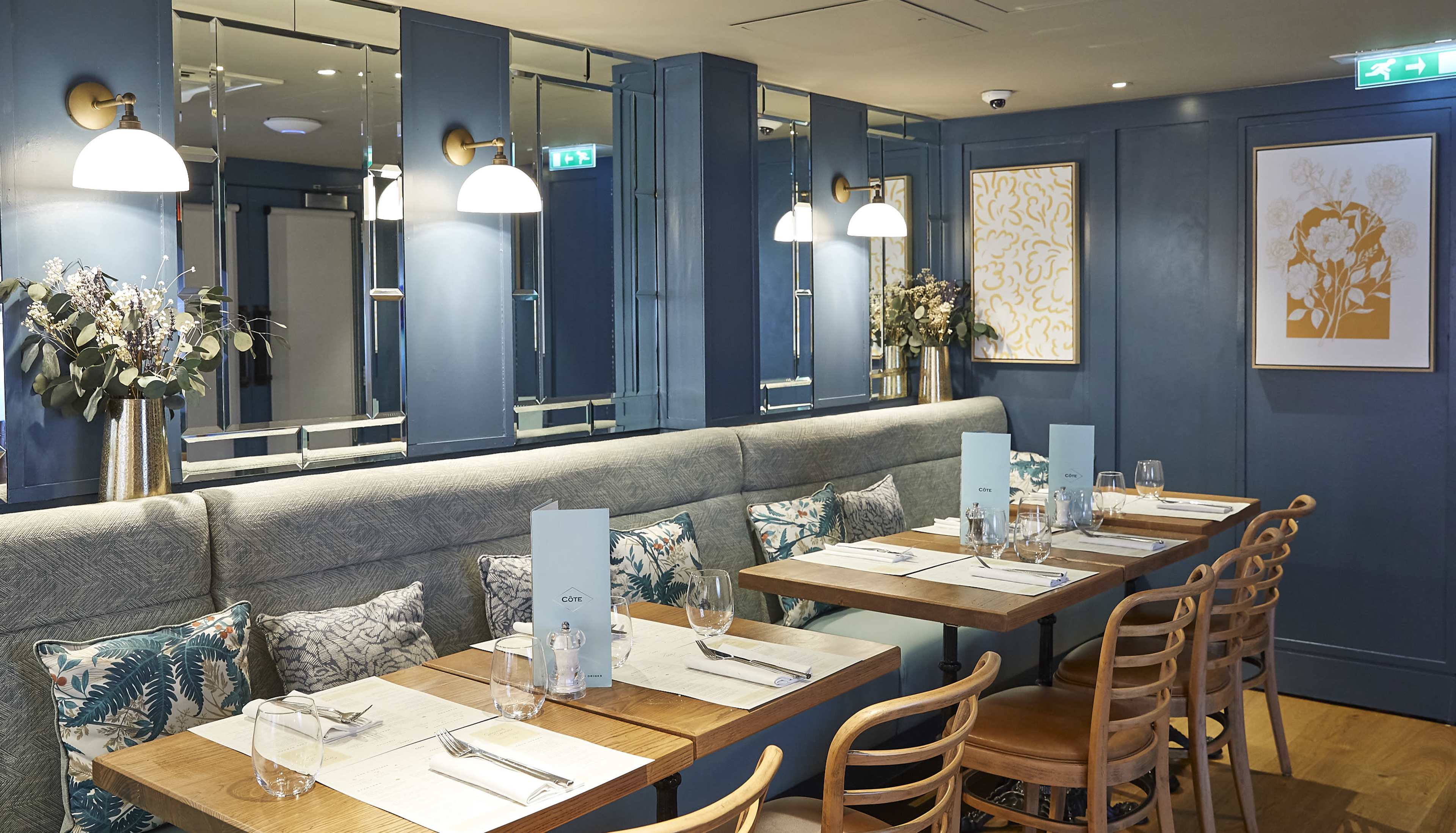 The image shows a stylish restaurant interior with teal walls, wooden tables, and upholstered seating, complemented by decorative lighting and floral arrangements.