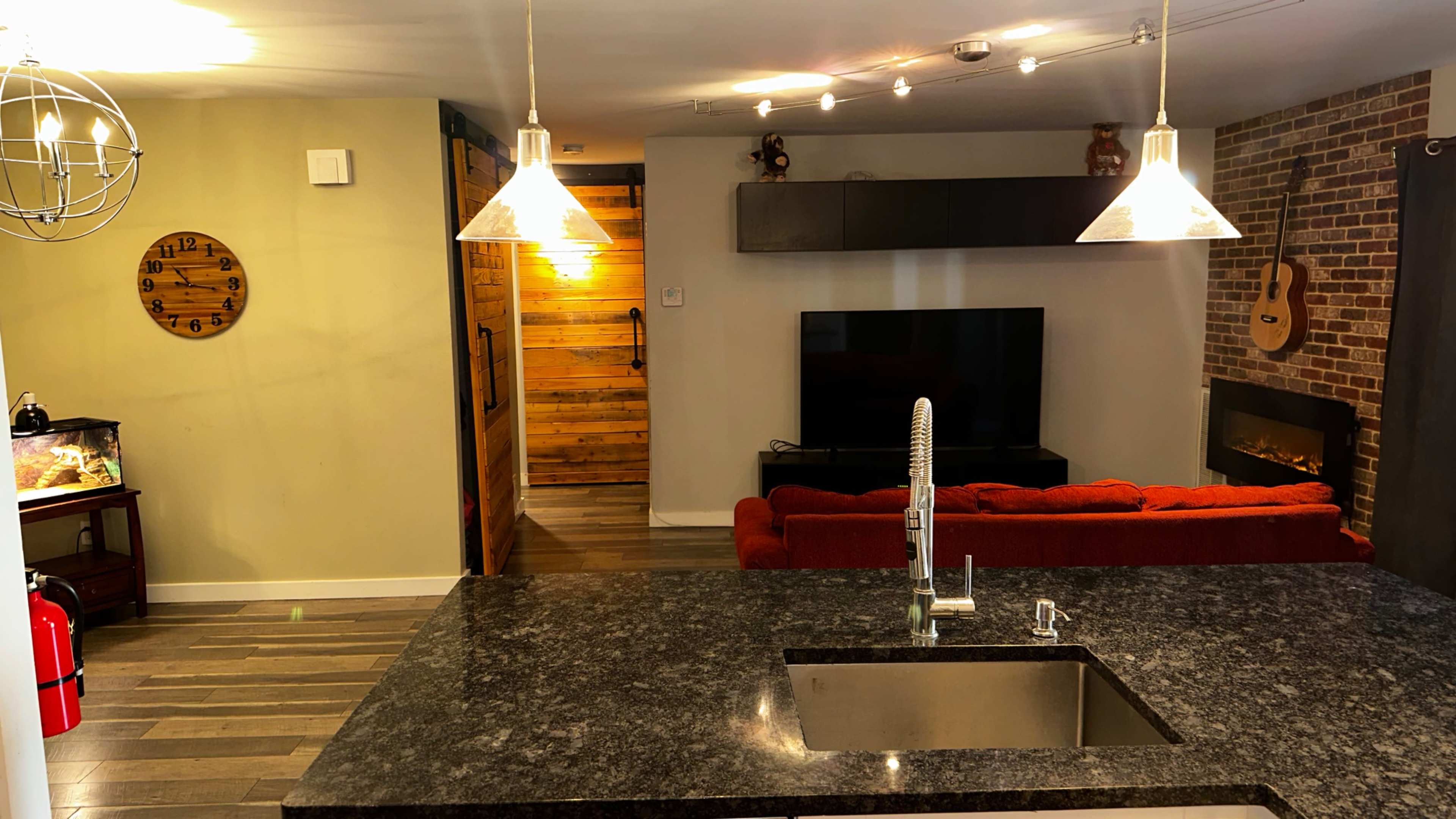 A modern kitchen with a dark granite countertop, a dining area in the foreground, and a living room featuring a television and a guitar hanging on the wall.