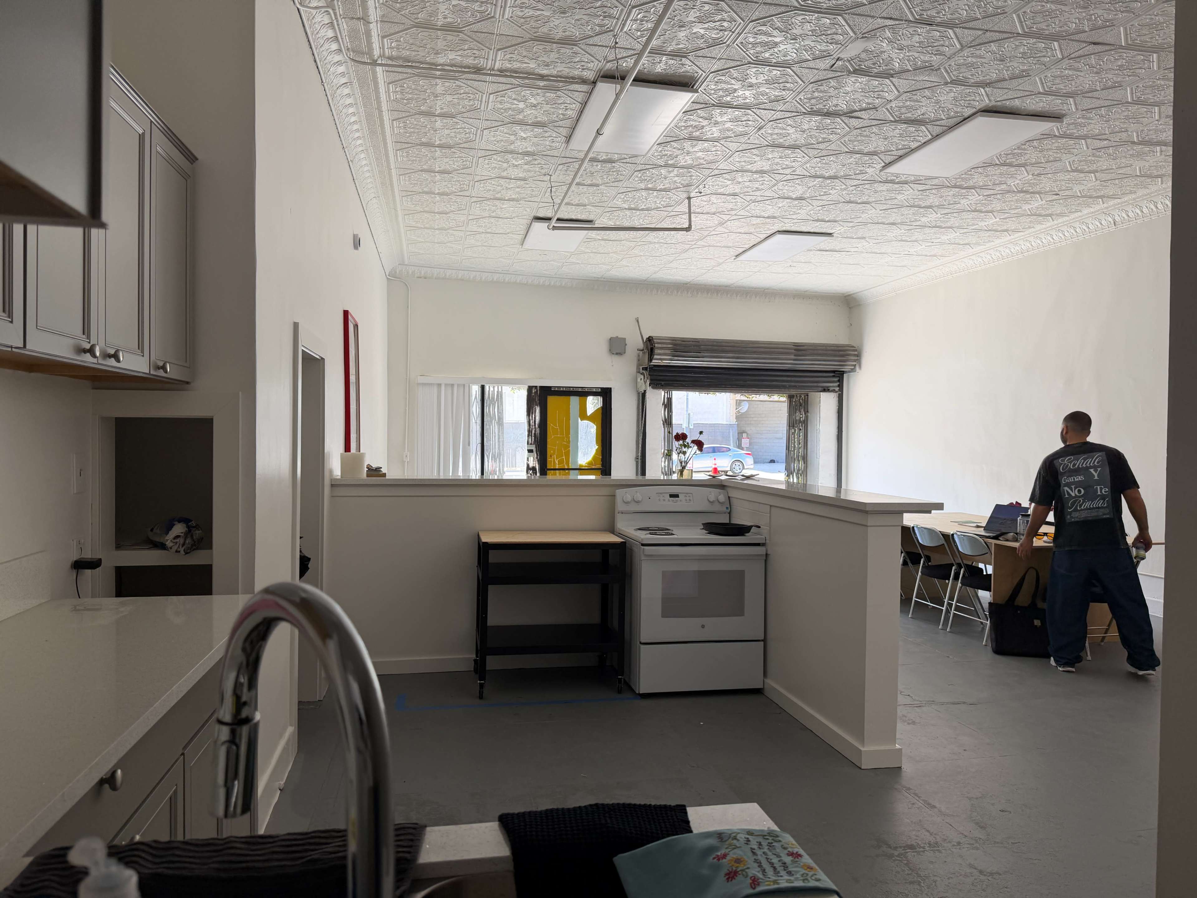 The image shows a spacious, well-lit kitchen area connected to a dining area with a man standing at a table in the background.