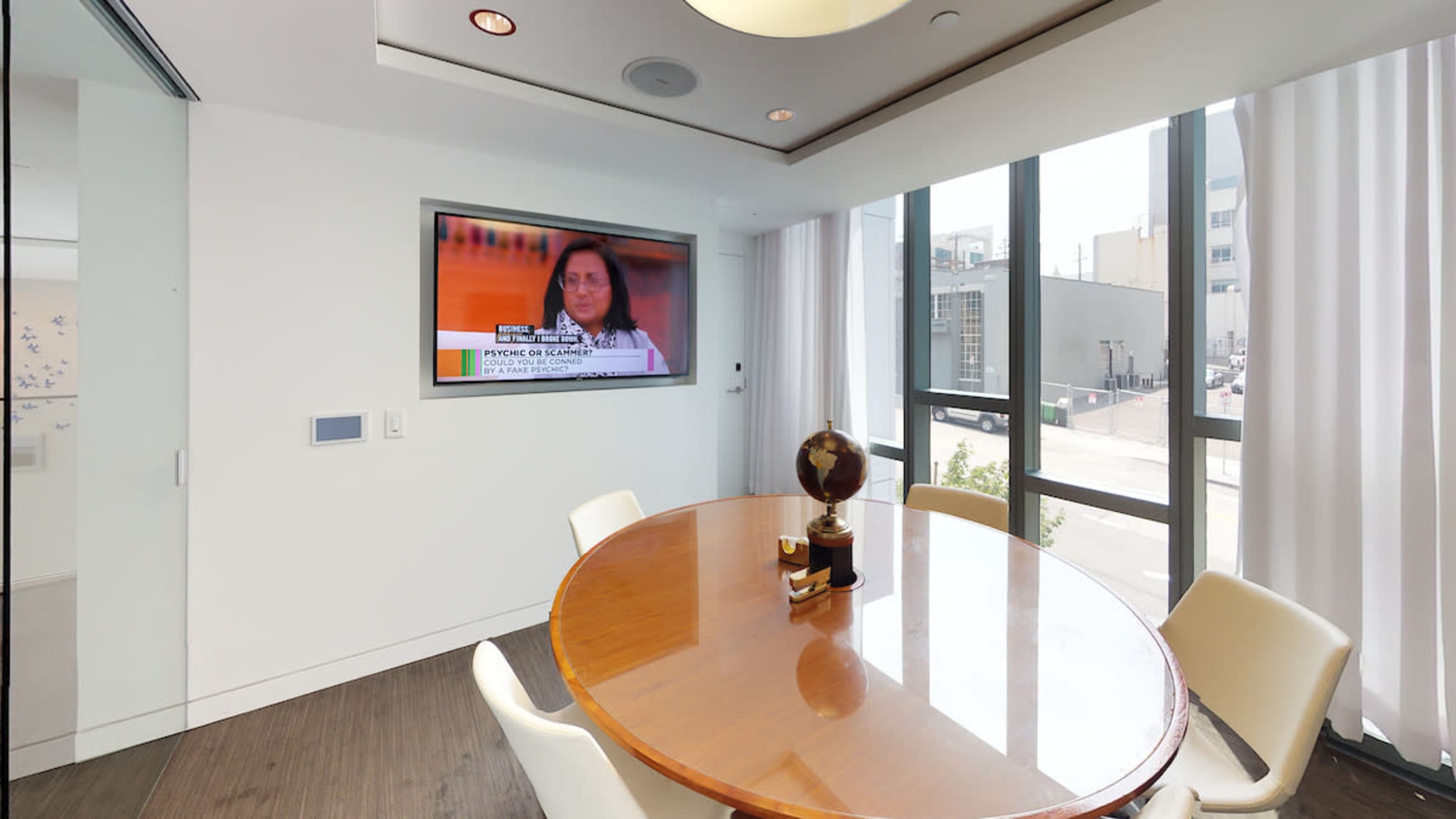 A round wooden table with chairs is seen in a well-lit room featuring a large television displaying a news segment.