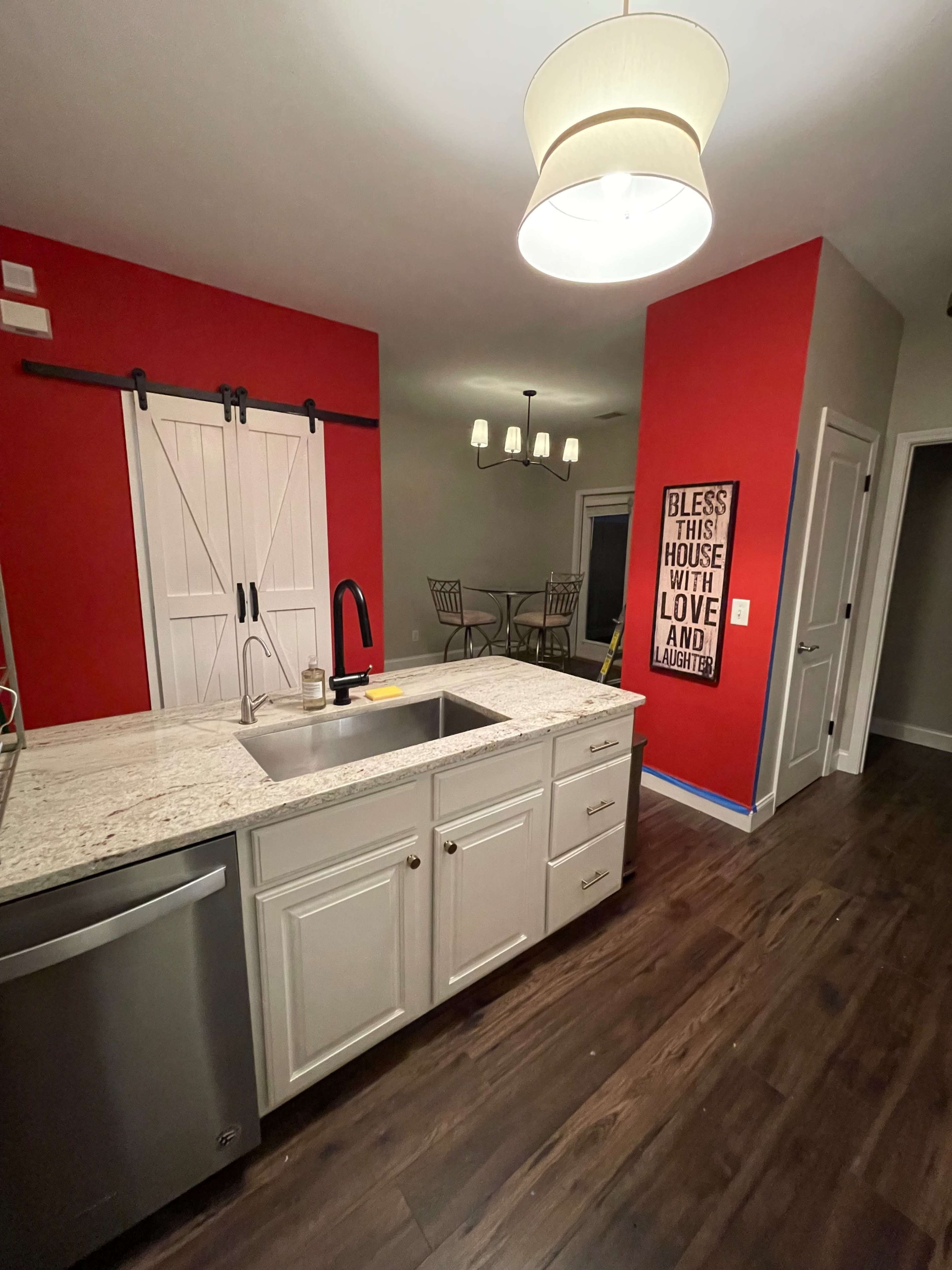 A kitchen with a granite countertop, a kitchen sink, and a red accent wall featuring a sliding barn door, with a dining area visible in the background.