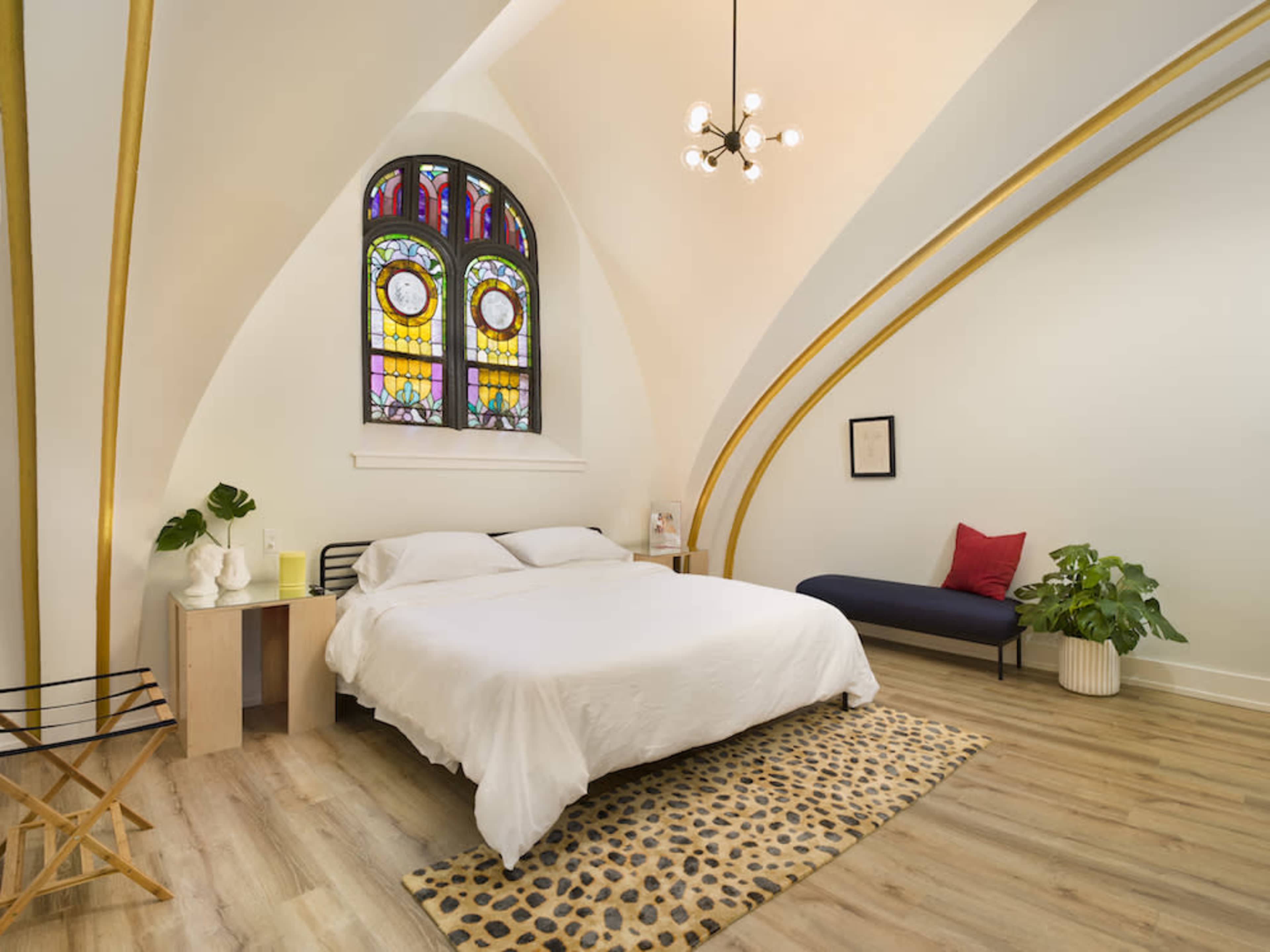 The image shows a spacious bedroom featuring a bed with white linens, a large stained glass window, a cozy seating area, and modern decor.