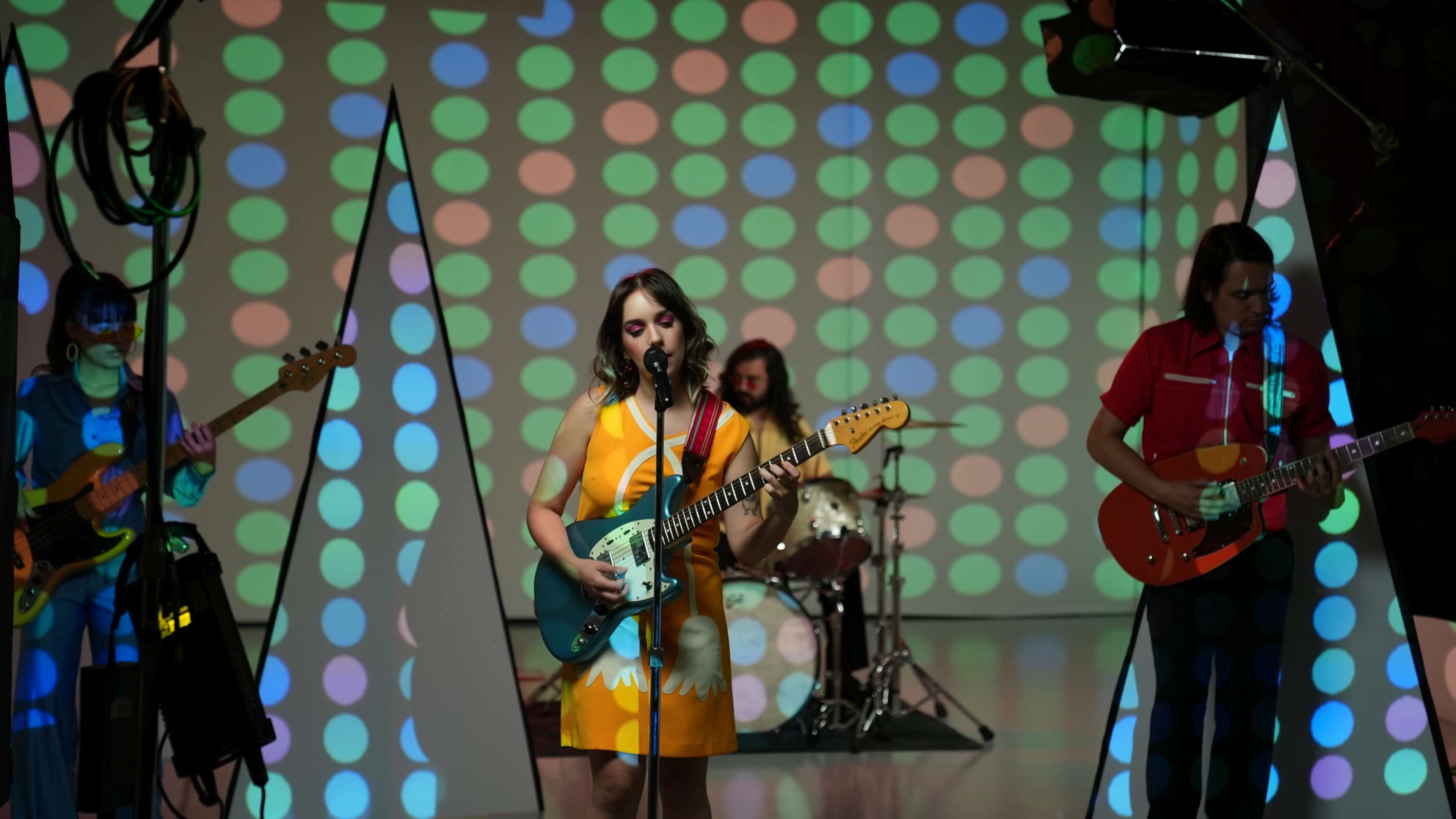 A band performs on stage with colorful polka dot projections on a backdrop, featuring a singer with an electric guitar and musicians playing drums and bass.