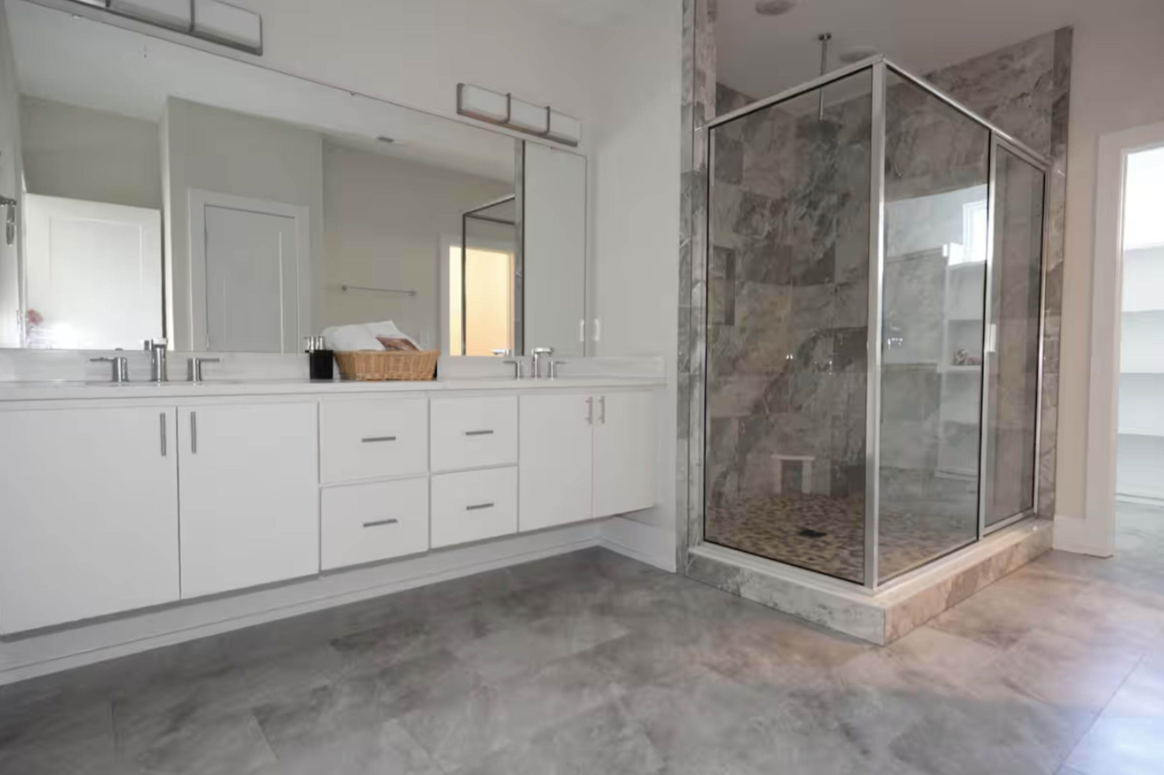 The image shows a modern bathroom featuring a large walk-in shower with glass walls, a double vanity with storage, and tiled flooring.