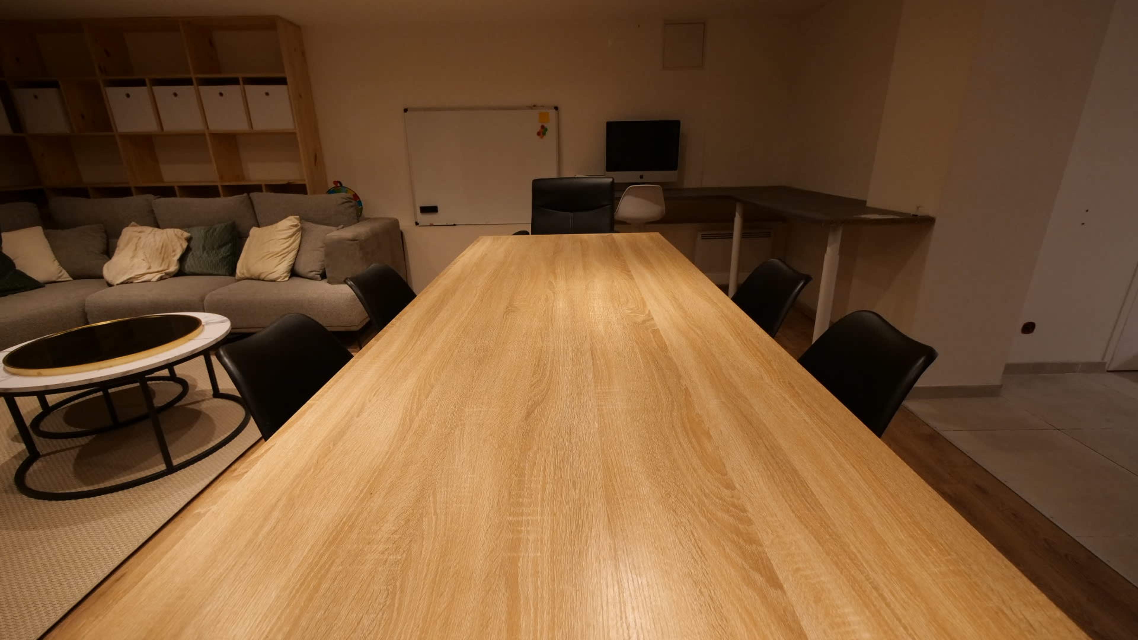 The image shows a large wooden dining table with several black chairs arranged around it in a room featuring a gray sofa and a whiteboard.