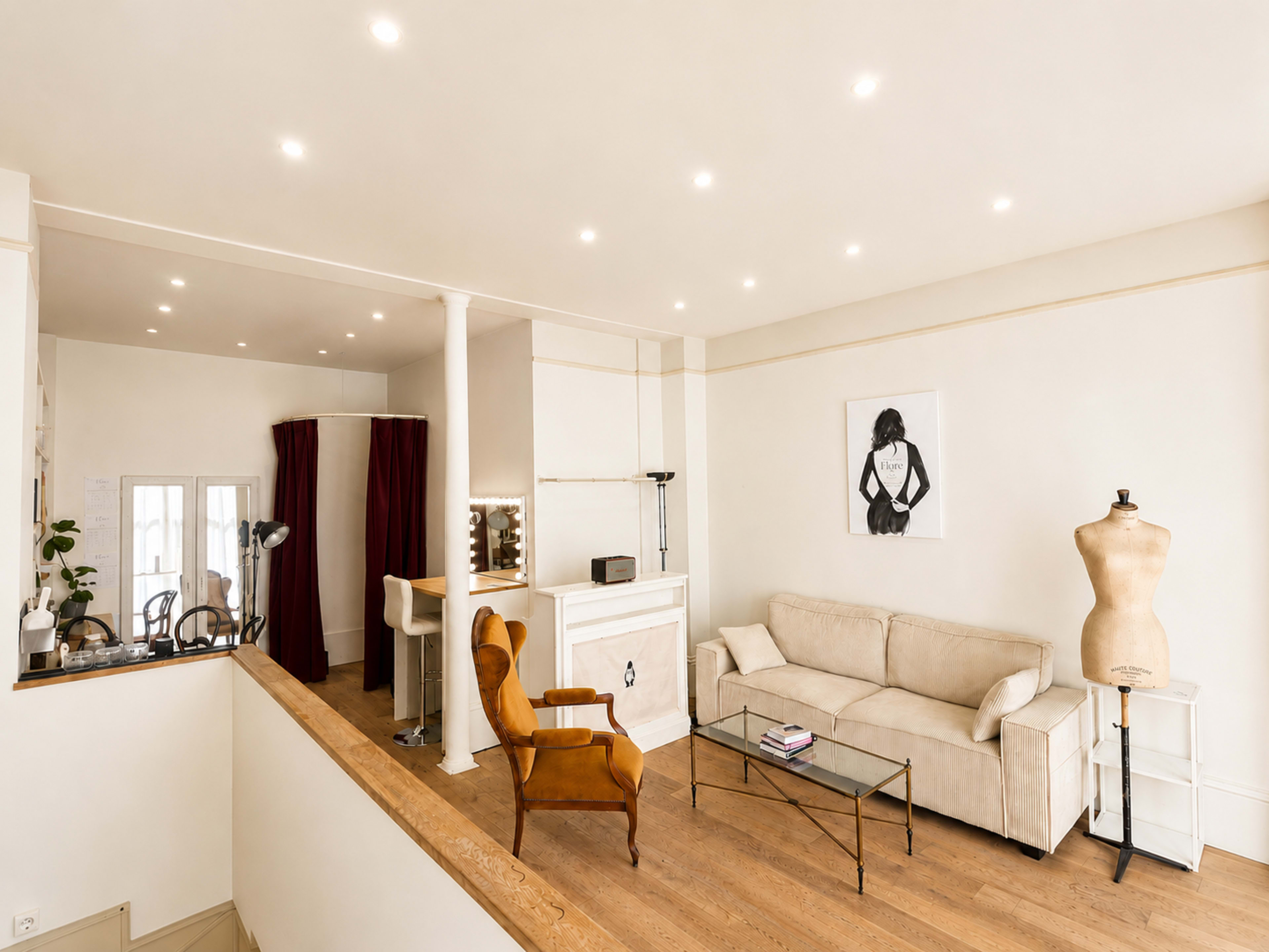 The image shows a bright, modern interior featuring a beige sofa, an orange chair, a glass coffee table, a mannequin, and a decorative poster on the wall.