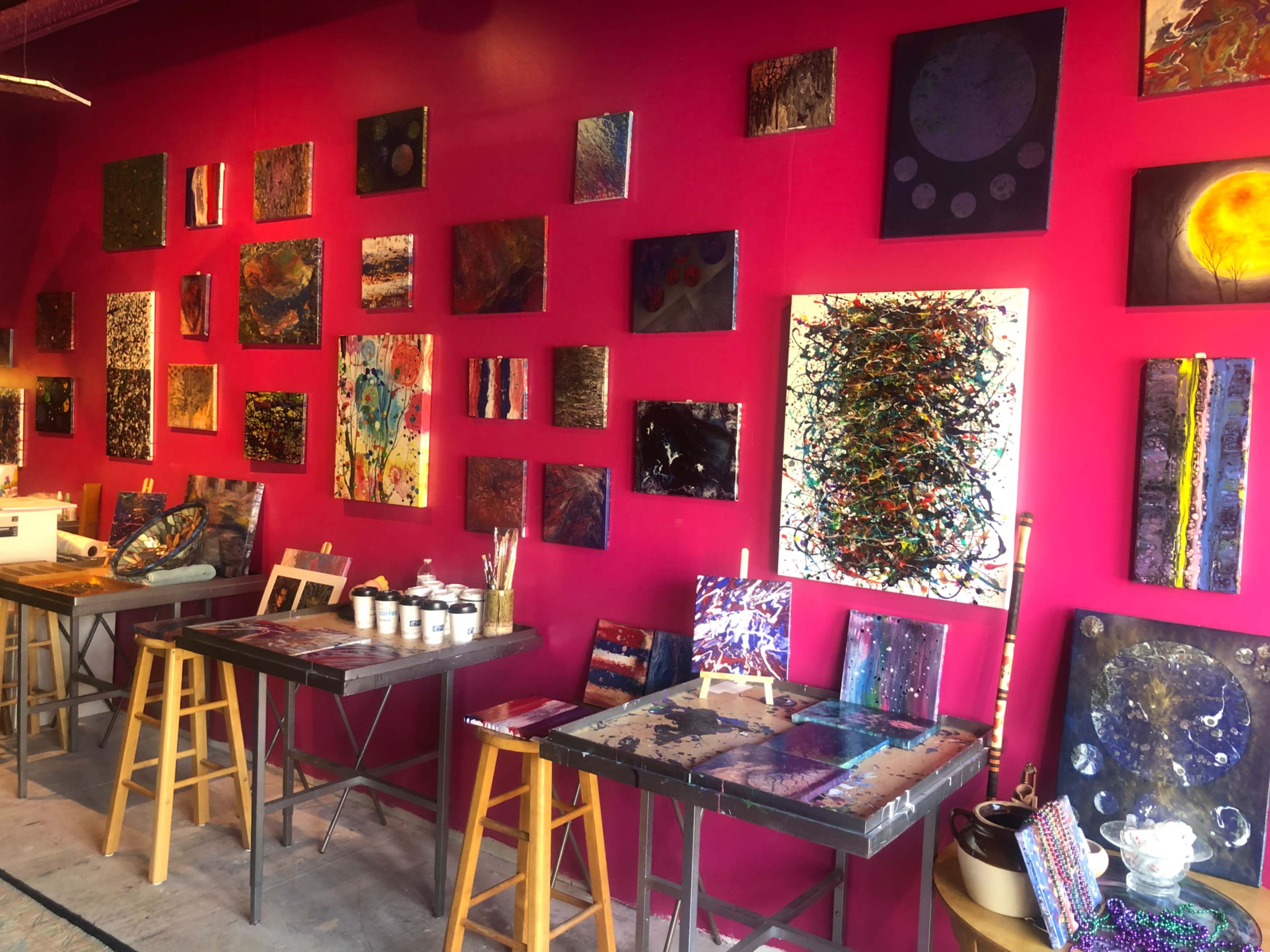 A vibrant art studio with a pink wall decorated with various paintings, and two tables holding art supplies and additional artworks.
