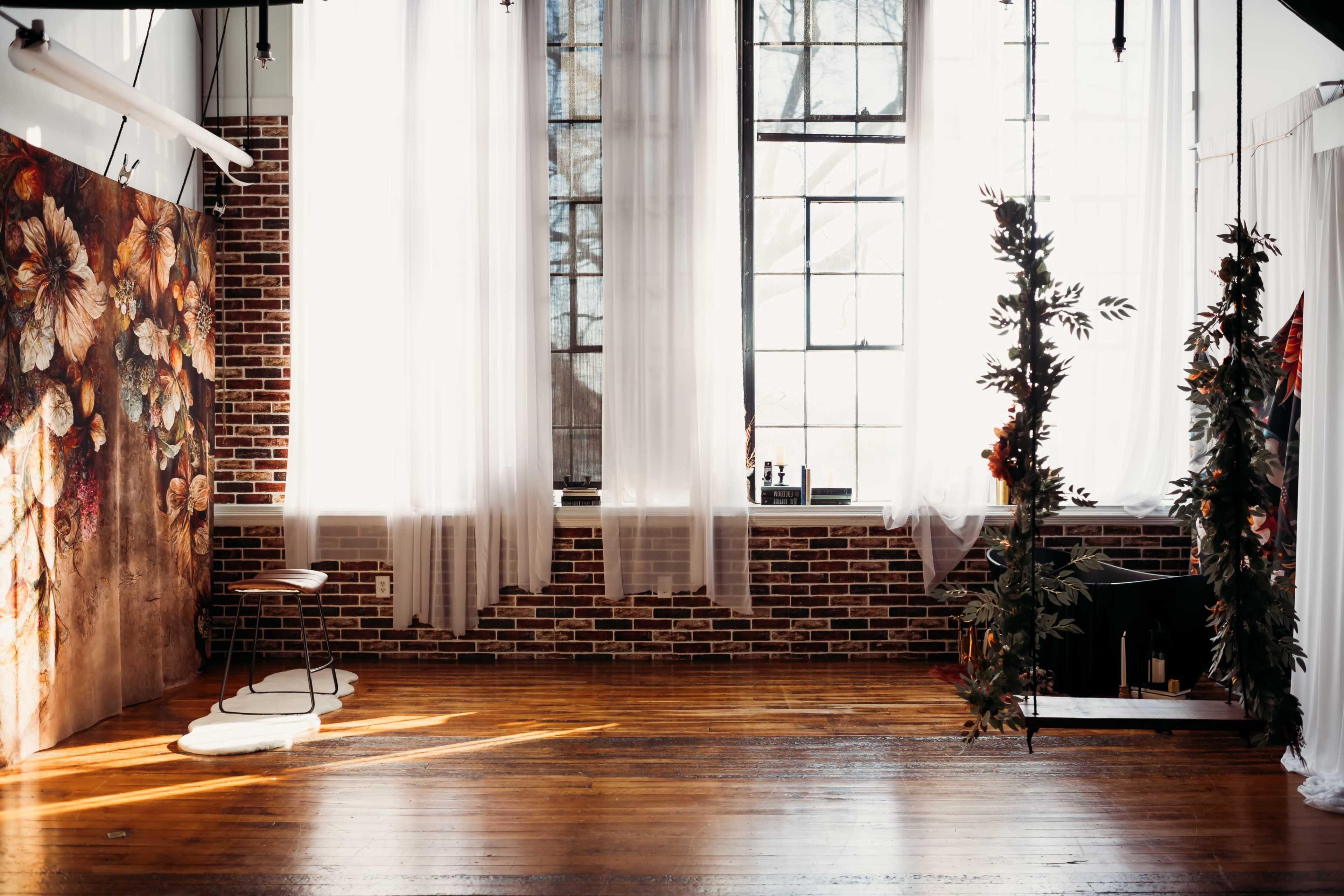 A spacious interior featuring large windows with sheer curtains, exposed brick walls, and wooden floors, with a floral backdrop and a hanging plant display.