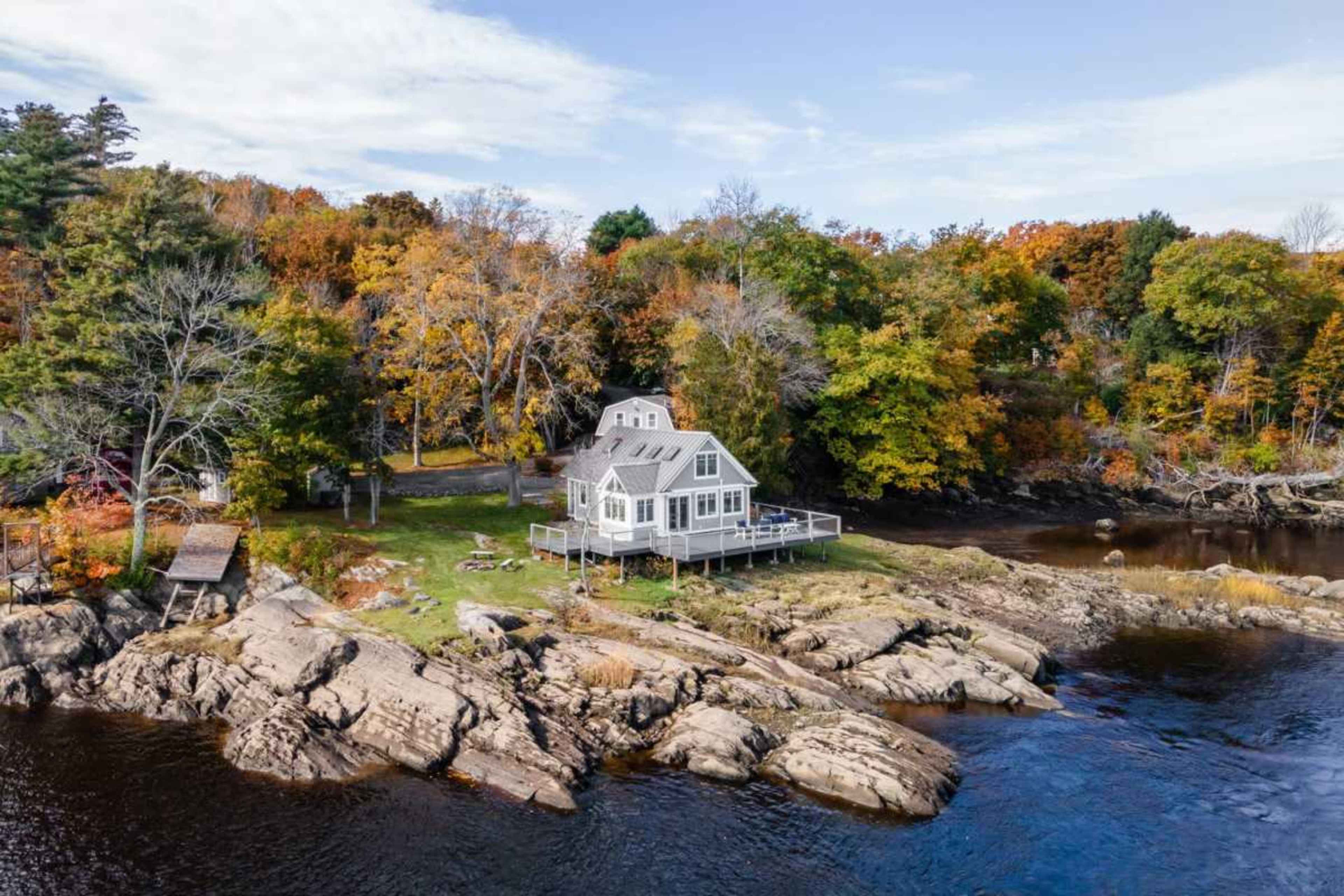 A white house with a porch is situated on rocky land near a river, surrounded by autumn-colored trees.