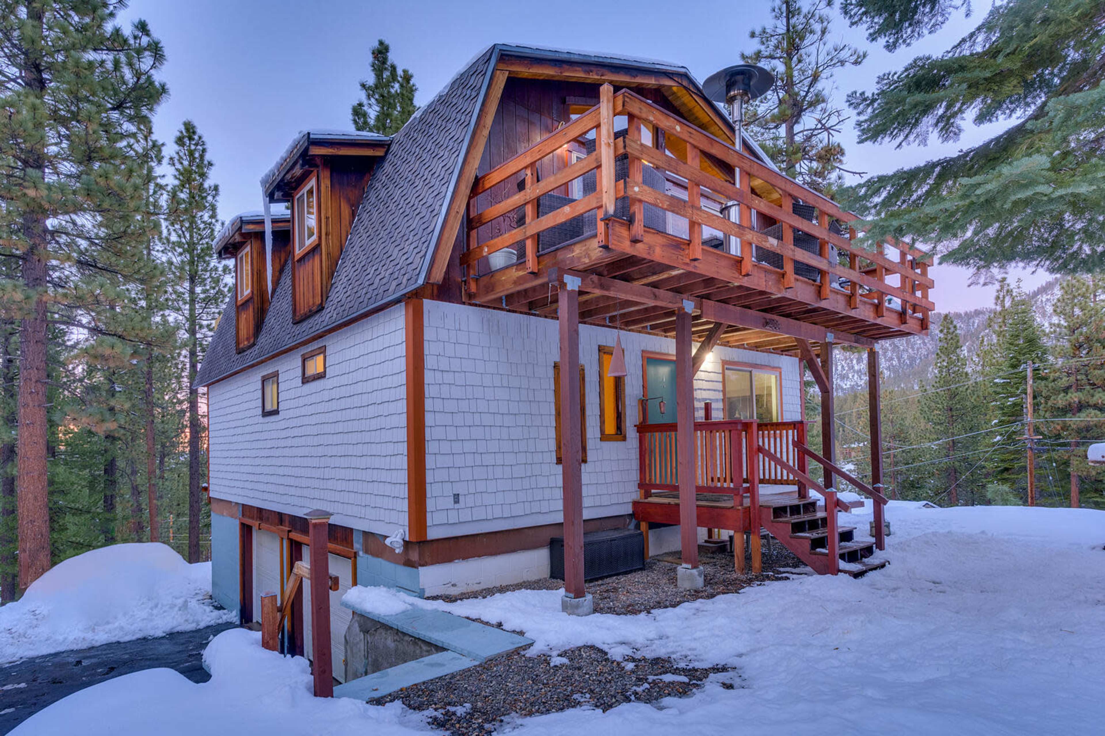 A two-story cabin with wooden accents and a balcony is situated in a snowy forested area.