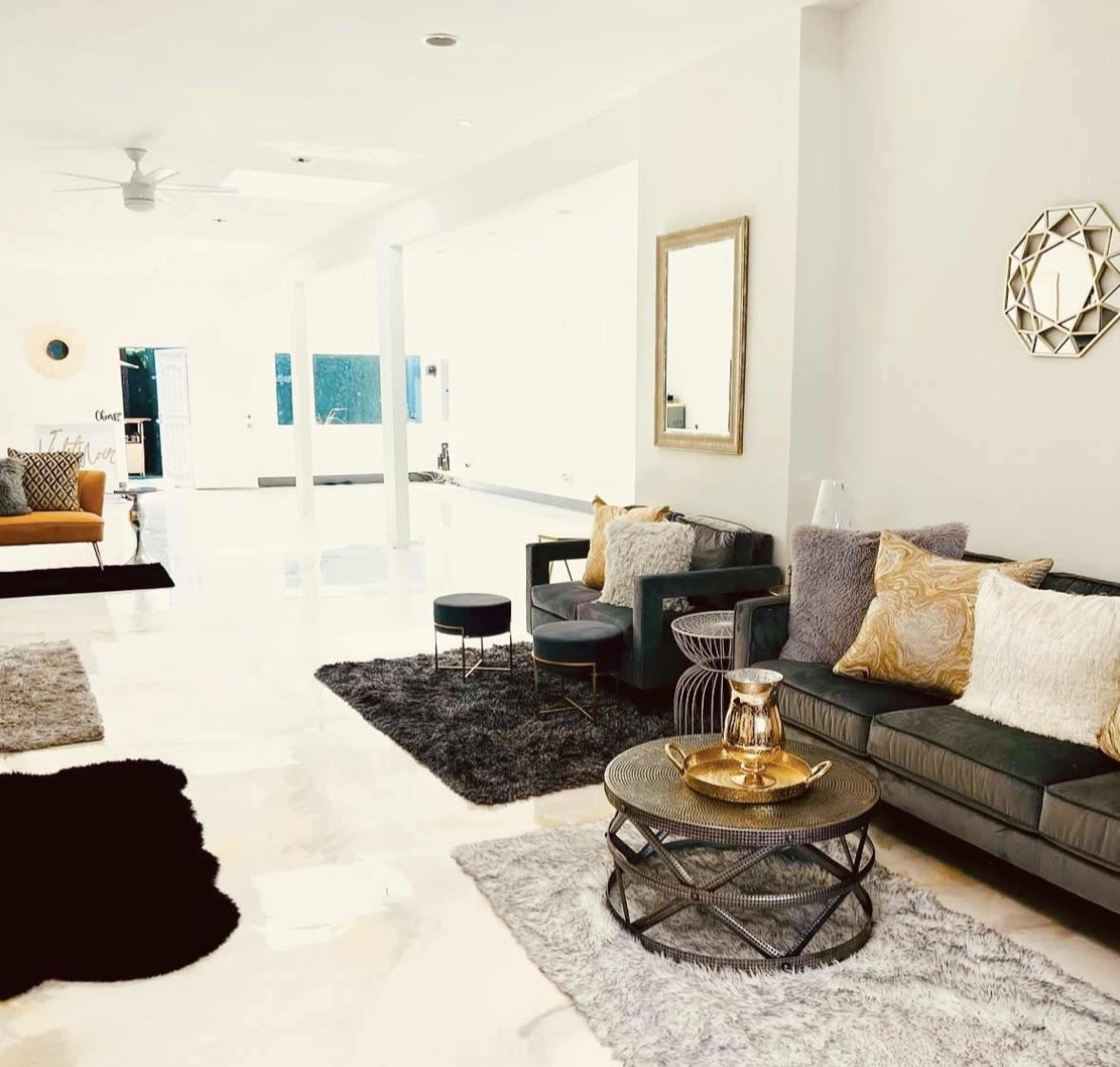The image shows a contemporary living room with polished marble floors, a dark sofa adorned with cushions, a glass coffee table, and various decorative elements including mirrors and rugs.