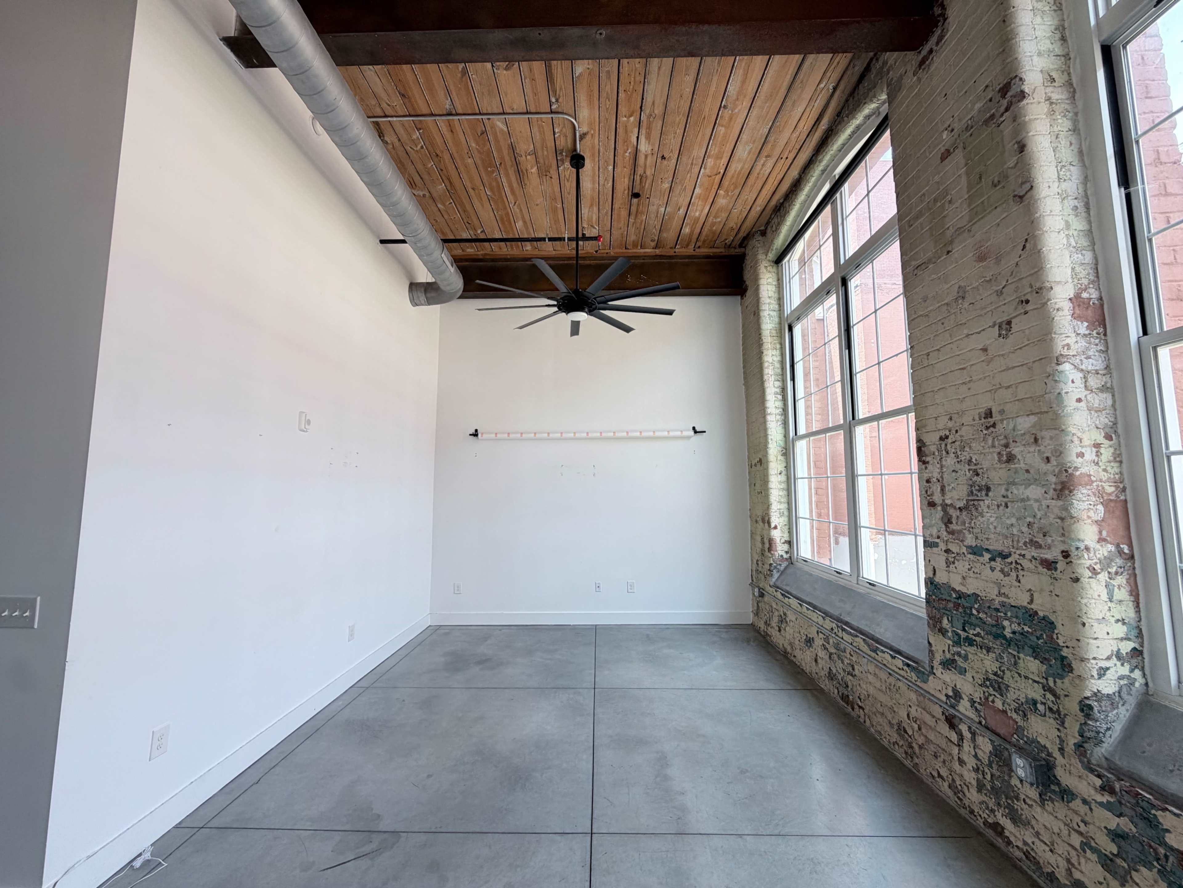 The image shows an empty room with exposed brick walls, a wooden ceiling, a ceiling fan, and large windows allowing natural light to enter.
