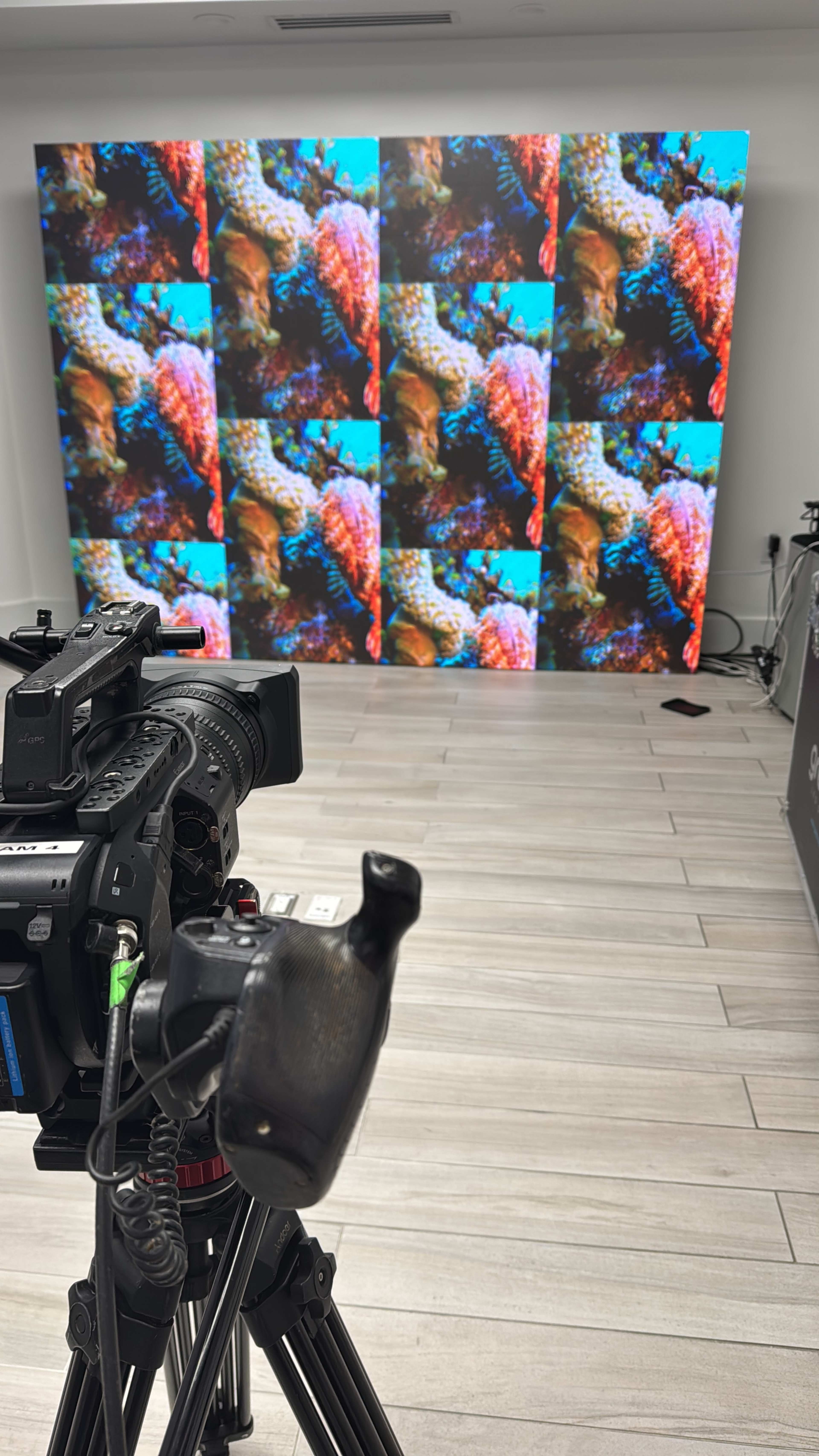 A camera is positioned in front of a large screen displaying colorful coral reef imagery.