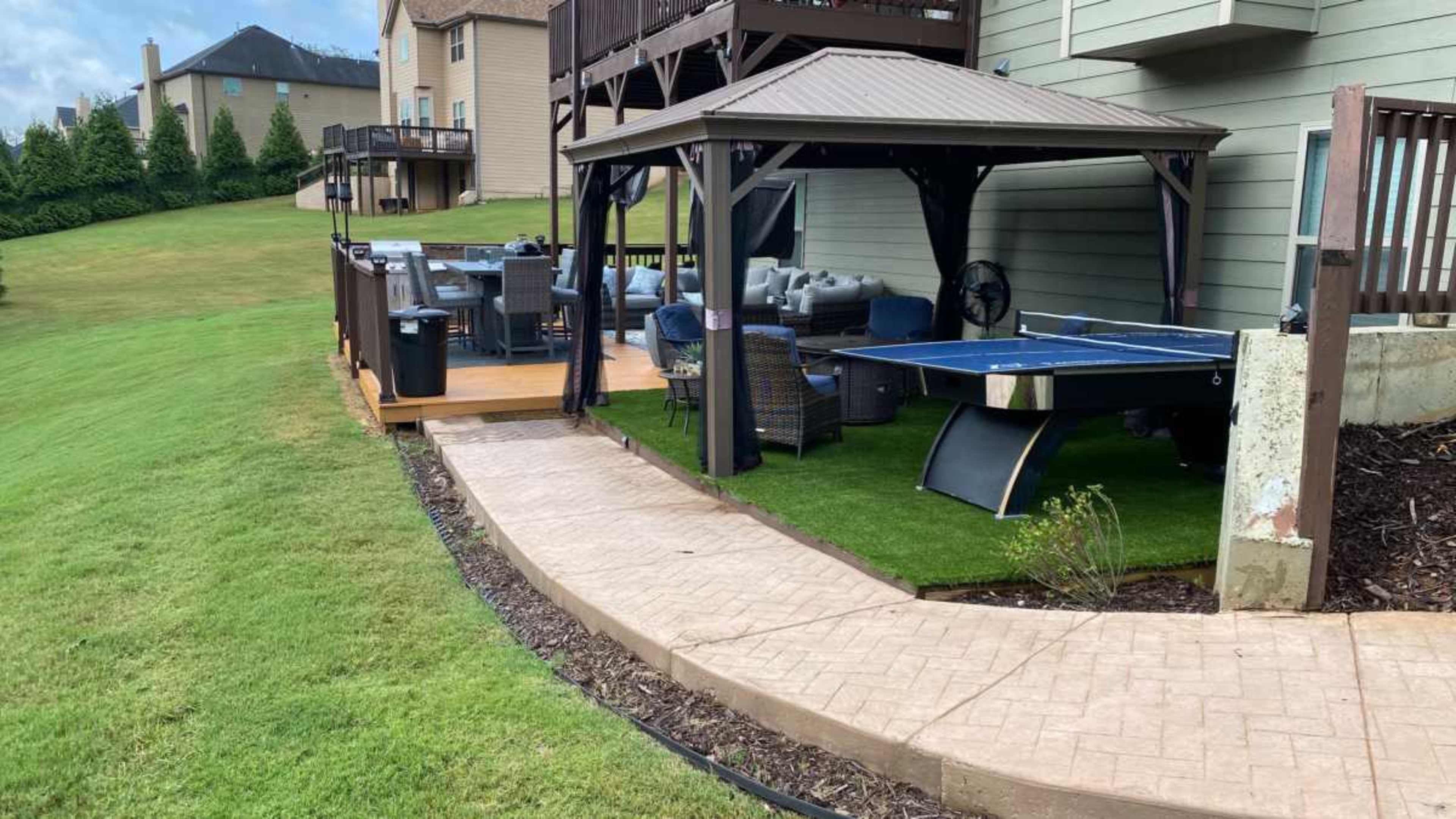 A backyard with a paved walkway leading to a covered patio area featuring outdoor furniture and a ping pong table.