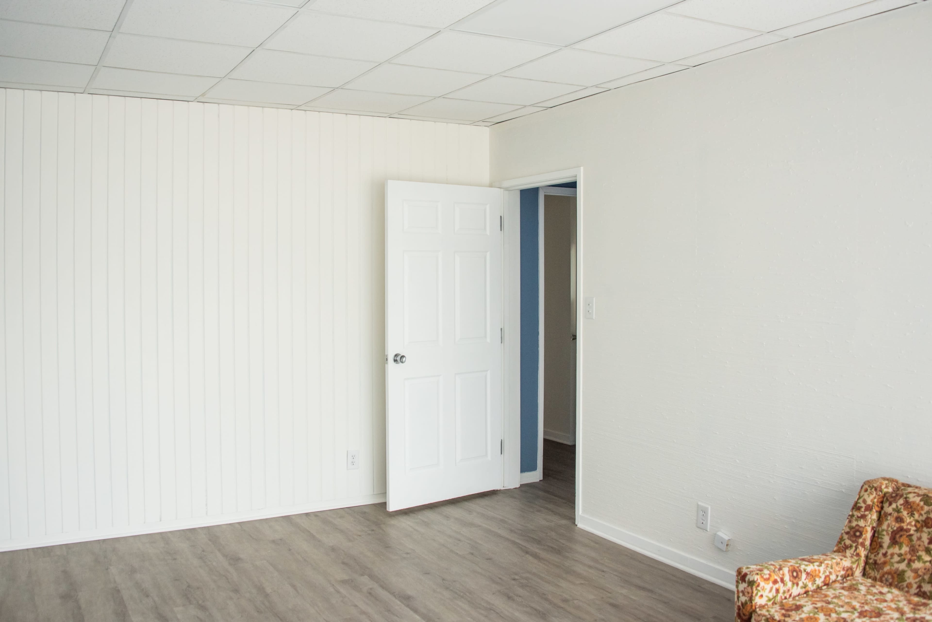 The image shows an empty room with a white paneled wall, a closed door on the left, and a patterned couch in the bottom right corner.