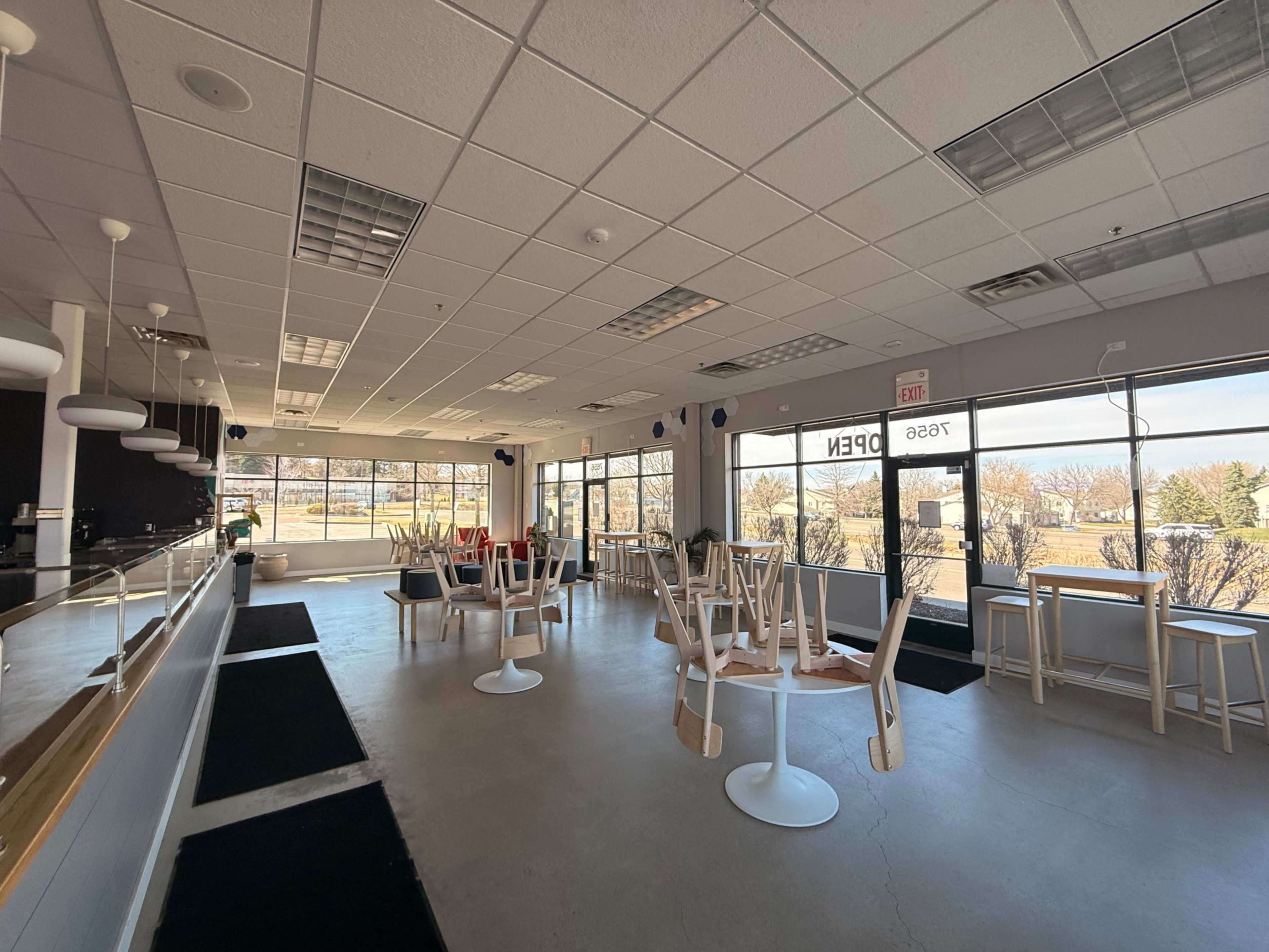 The image shows a brightly lit, empty café with several chairs stacked upside down on tables and large windows overlooking an outdoor view.