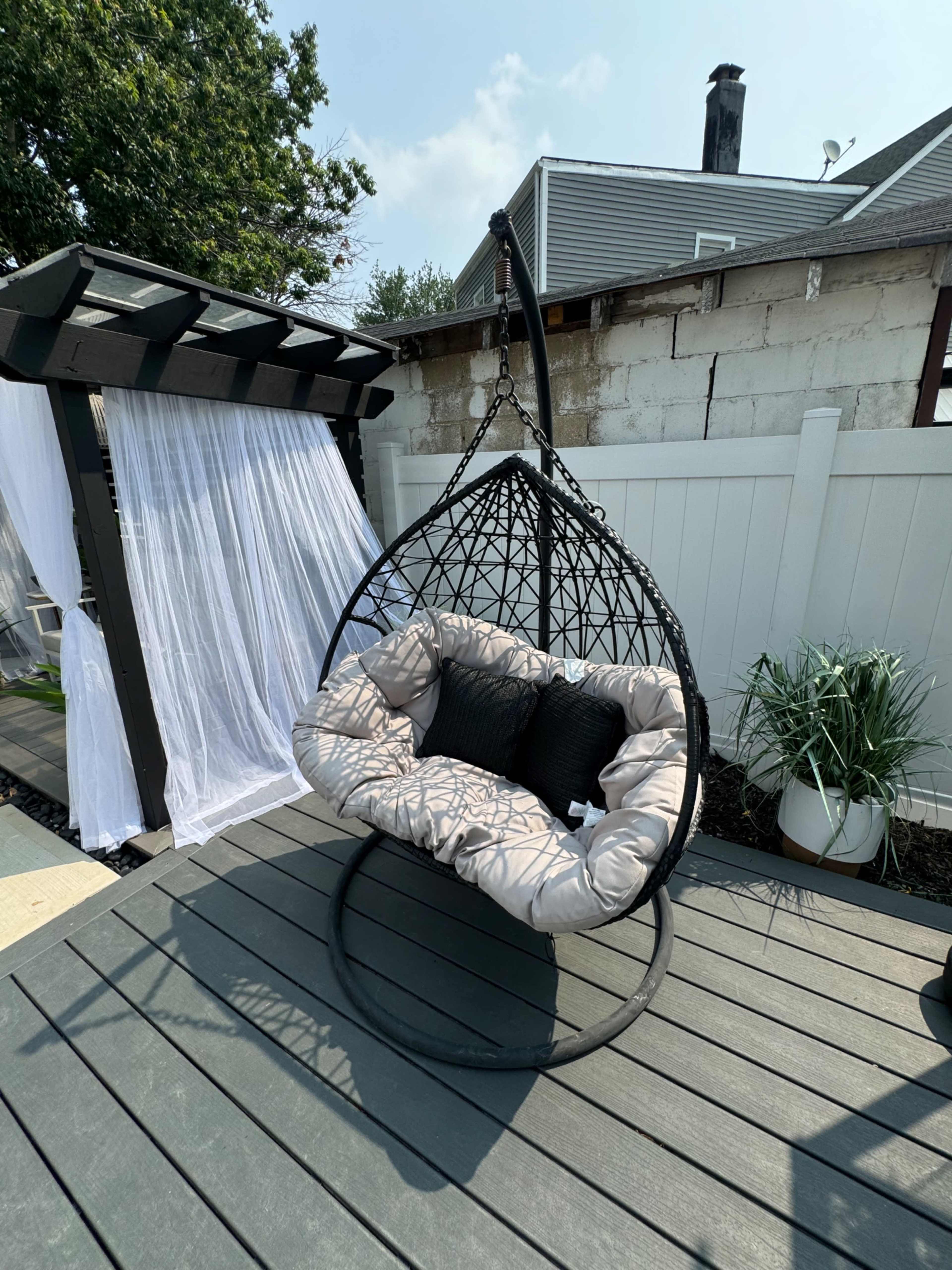 A hanging chair with a cushioned seat is positioned on a wooden deck next to a white privacy fence and a canopy structure.