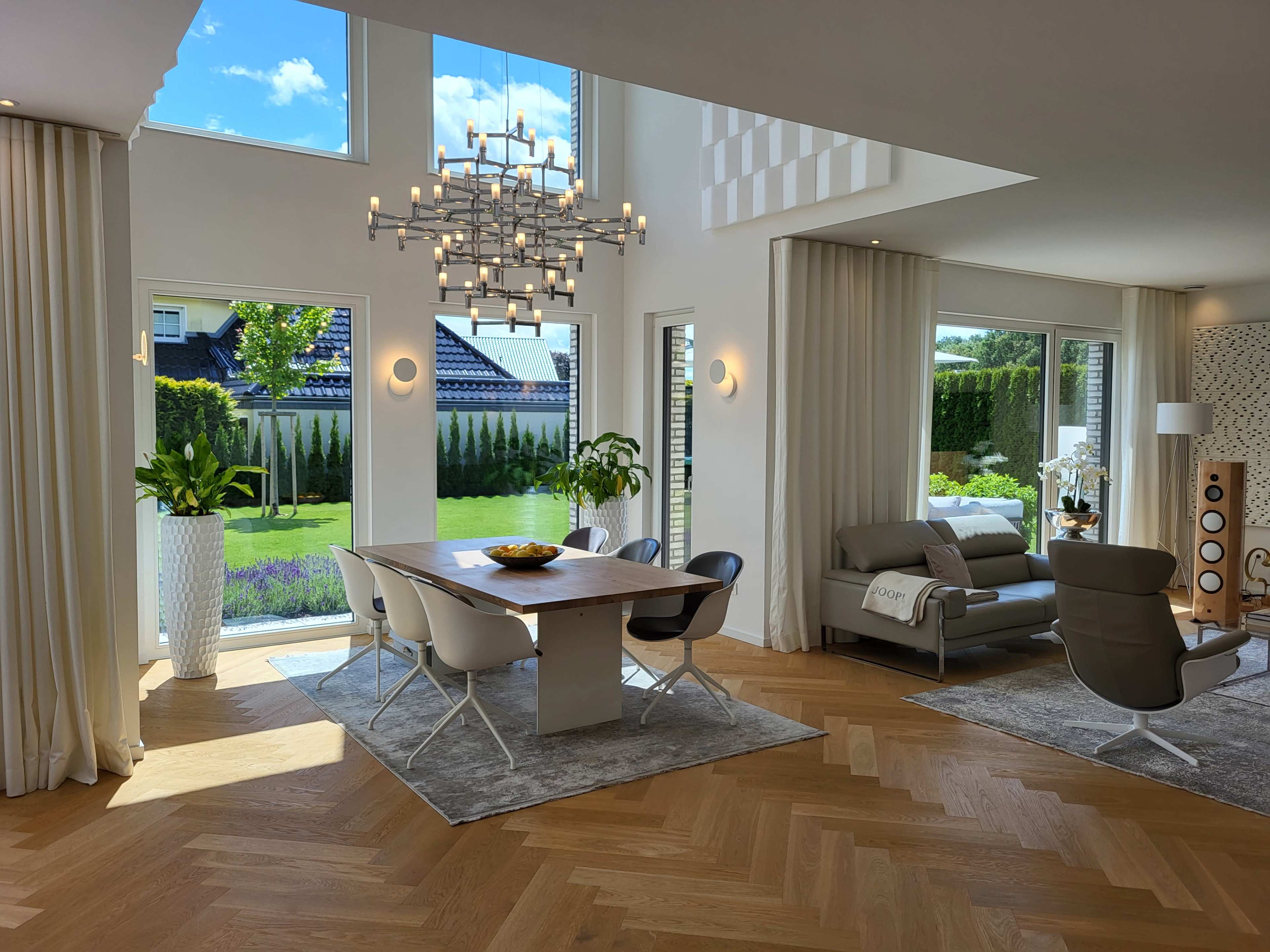 A modern living and dining area with large windows, a dining table, stylish chairs, and a contemporary sofa, surrounded by greenery.
