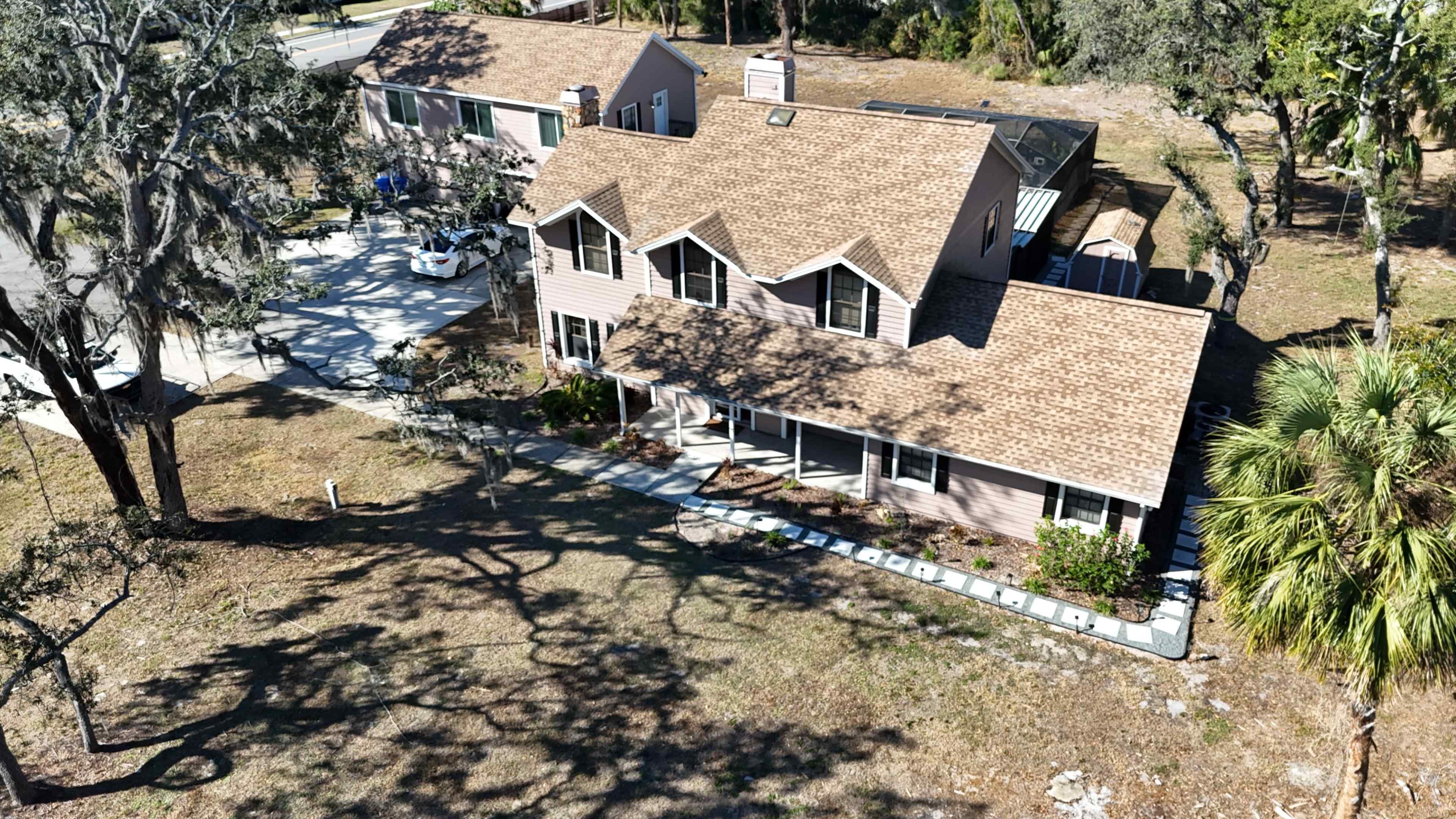 The image shows a large, two-story house with a brown shingled roof surrounded by trees and a clear driveway.