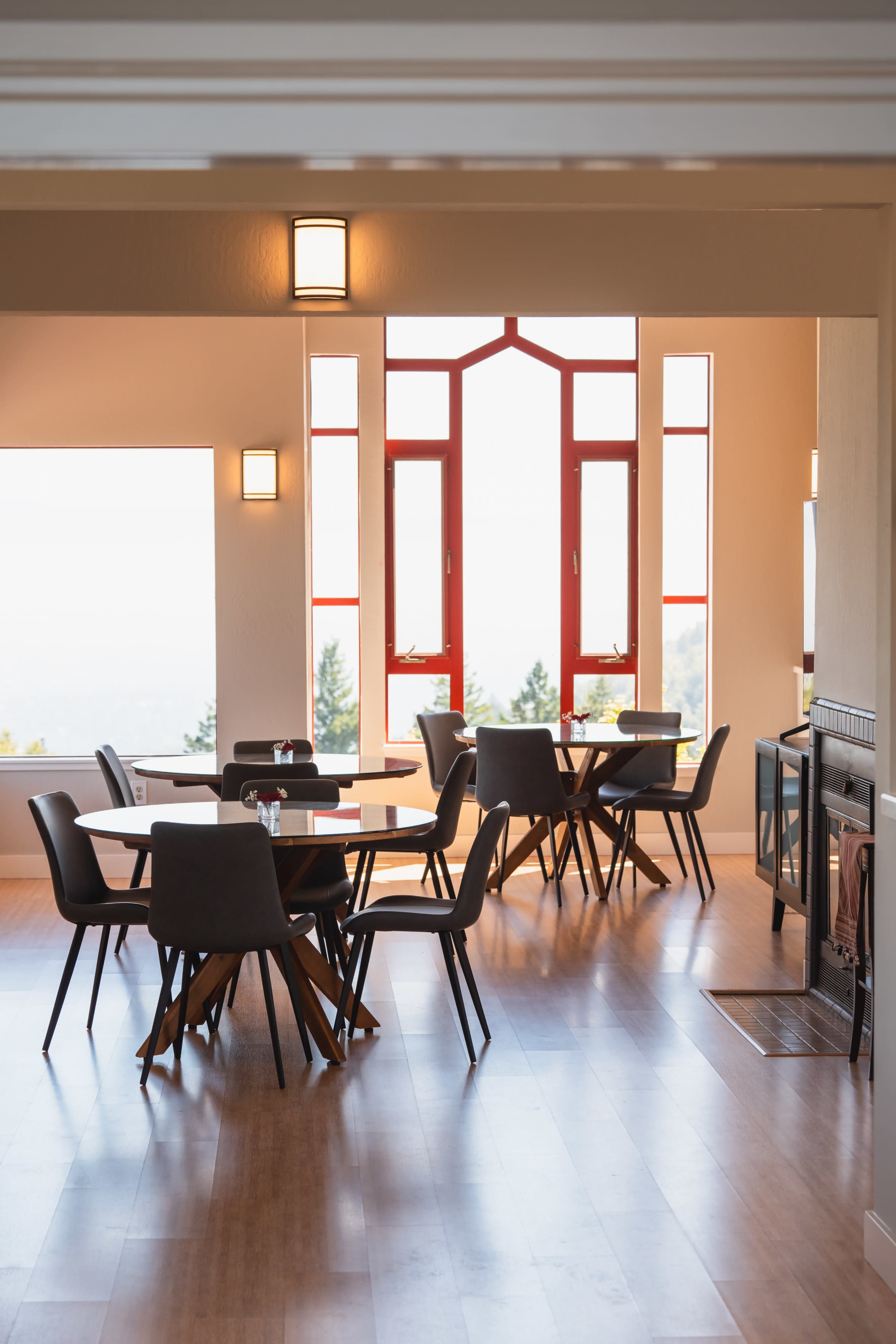 The image shows a modern dining area with several round tables, chairs, and large windows featuring red frames that offer a view.