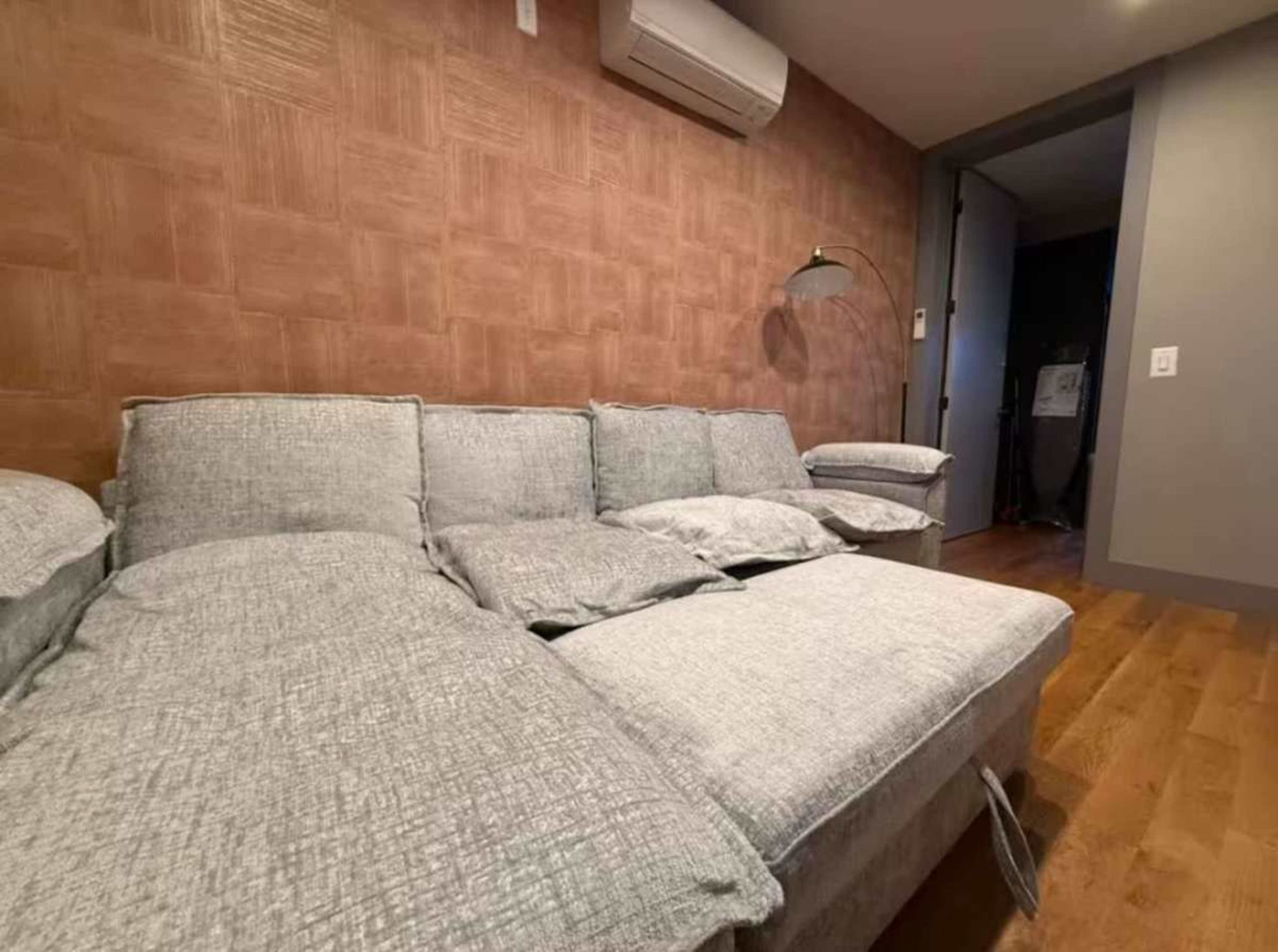 The image shows a modern living room with a light gray sectional sofa against a textured brown wall, accompanied by a floor lamp and an air conditioning unit.