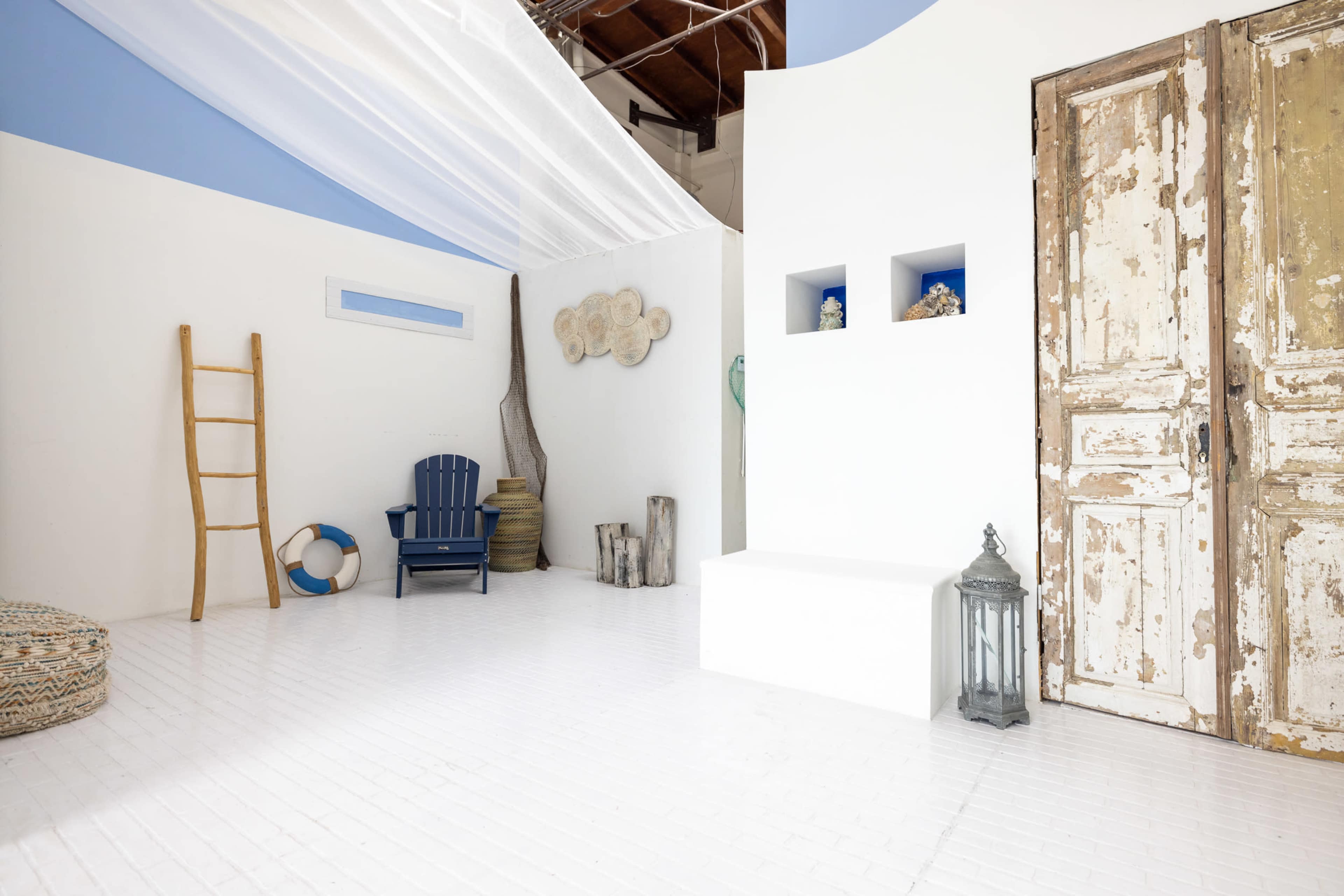 The image shows a bright, minimalist interior space with white walls, a blue ceiling, a large wooden door, and a mix of decorative elements including a ladder and a lantern.