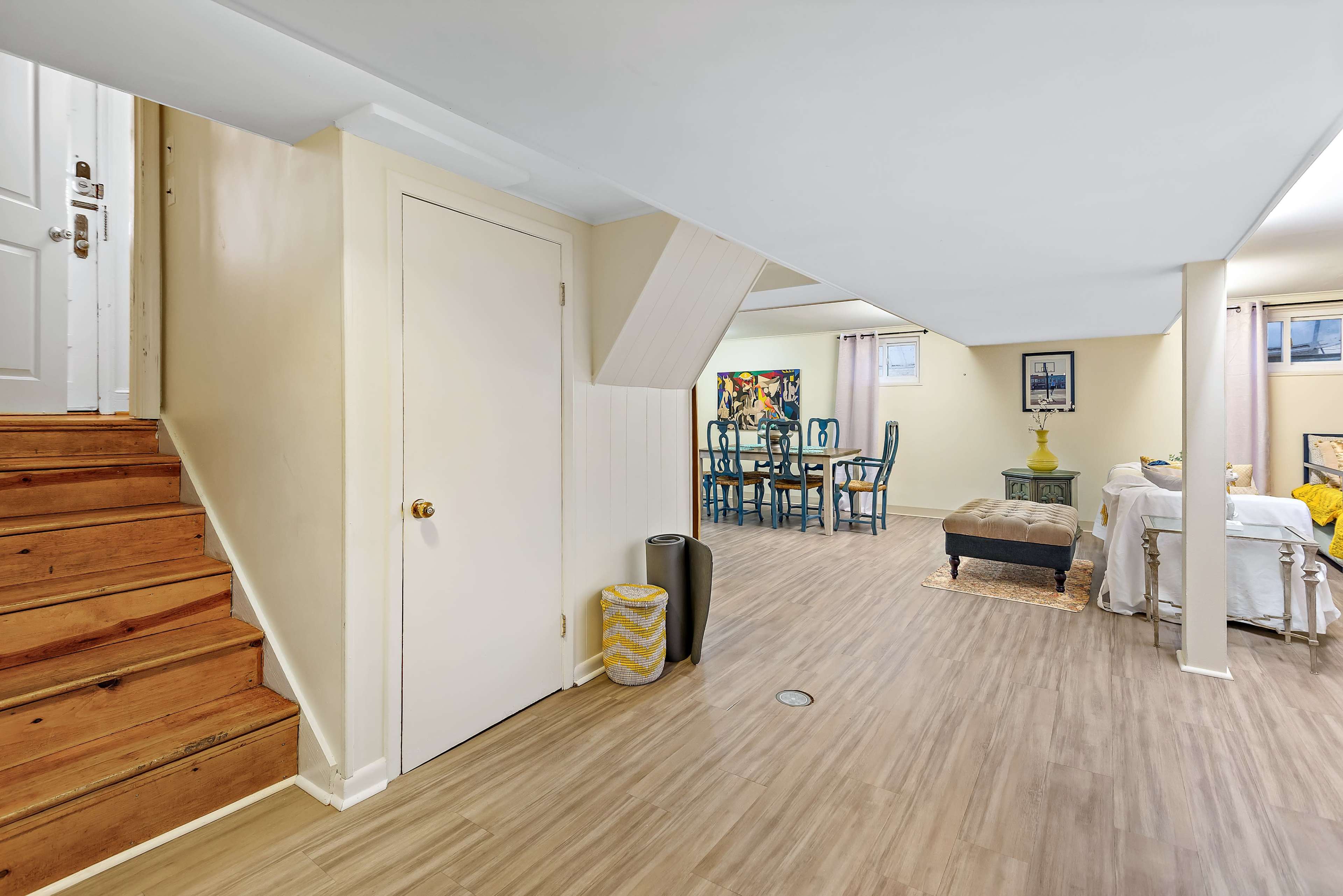 A basement living area featuring a staircase, a closed door, and a dining table with chairs in the background.