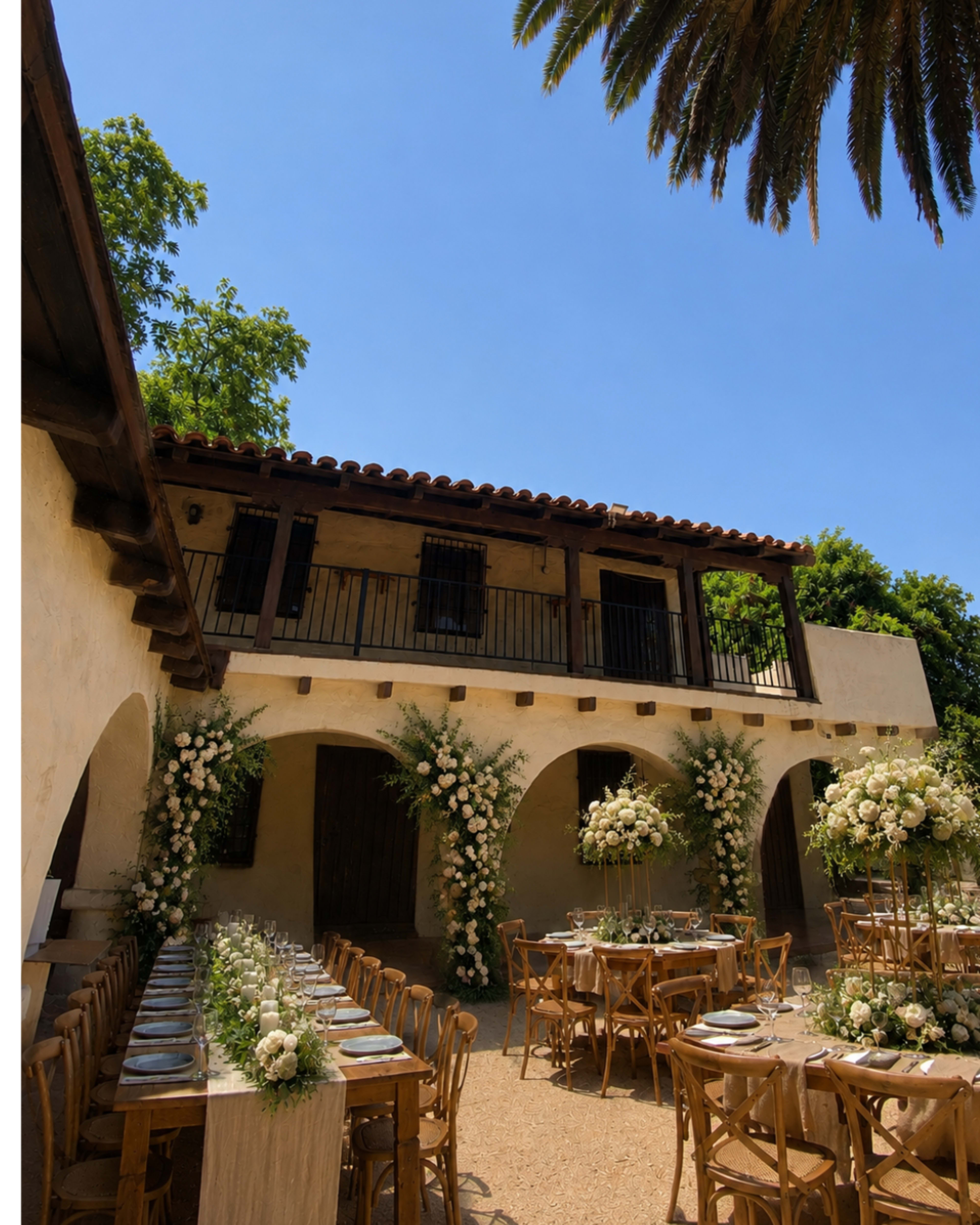 A rustic outdoor venue features wooden tables set for dining, surrounded by floral arrangements and a building with a tile roof under a clear blue sky.