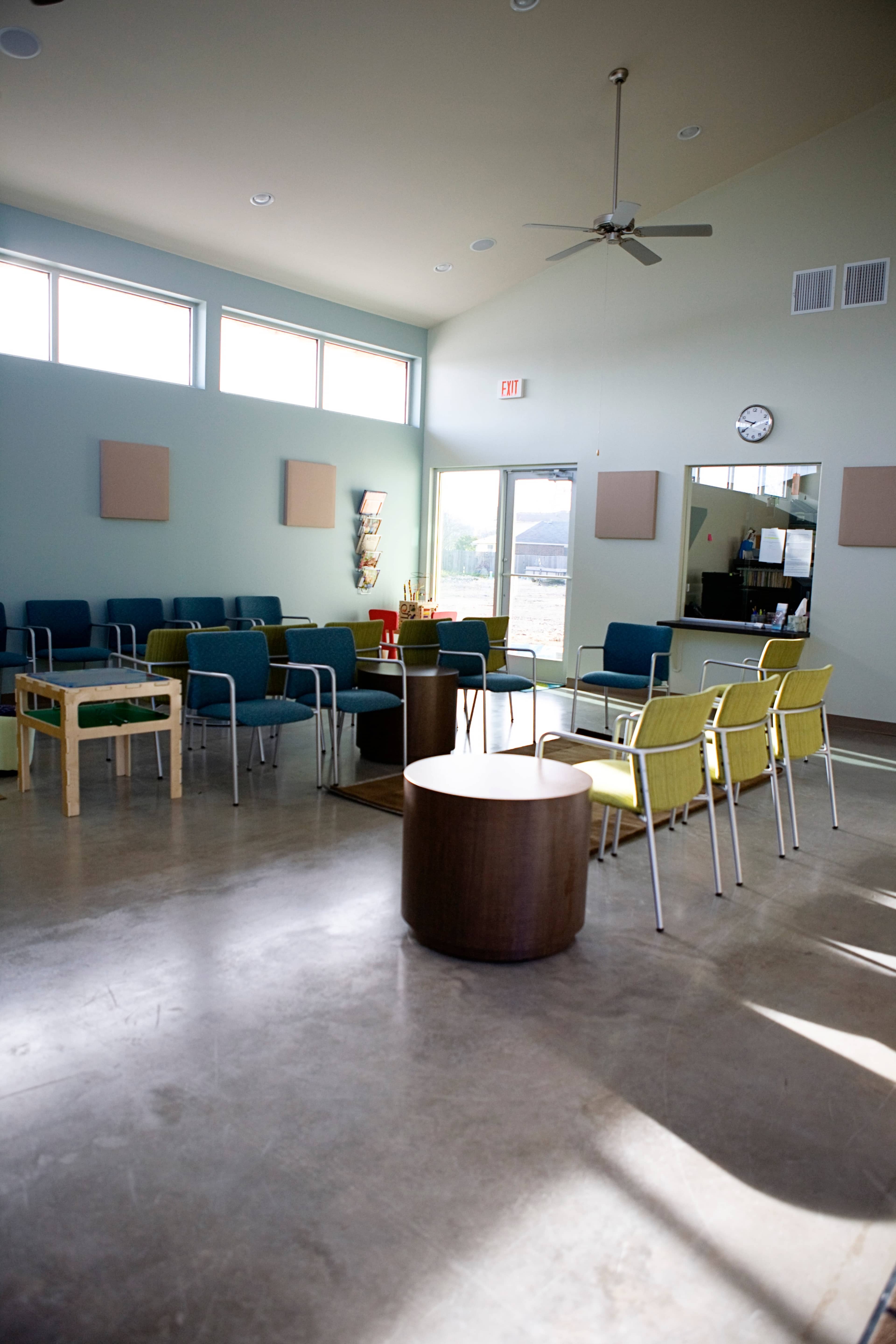 A spacious interior of a community room featuring various seating arrangements, large windows, and a kitchenette area.