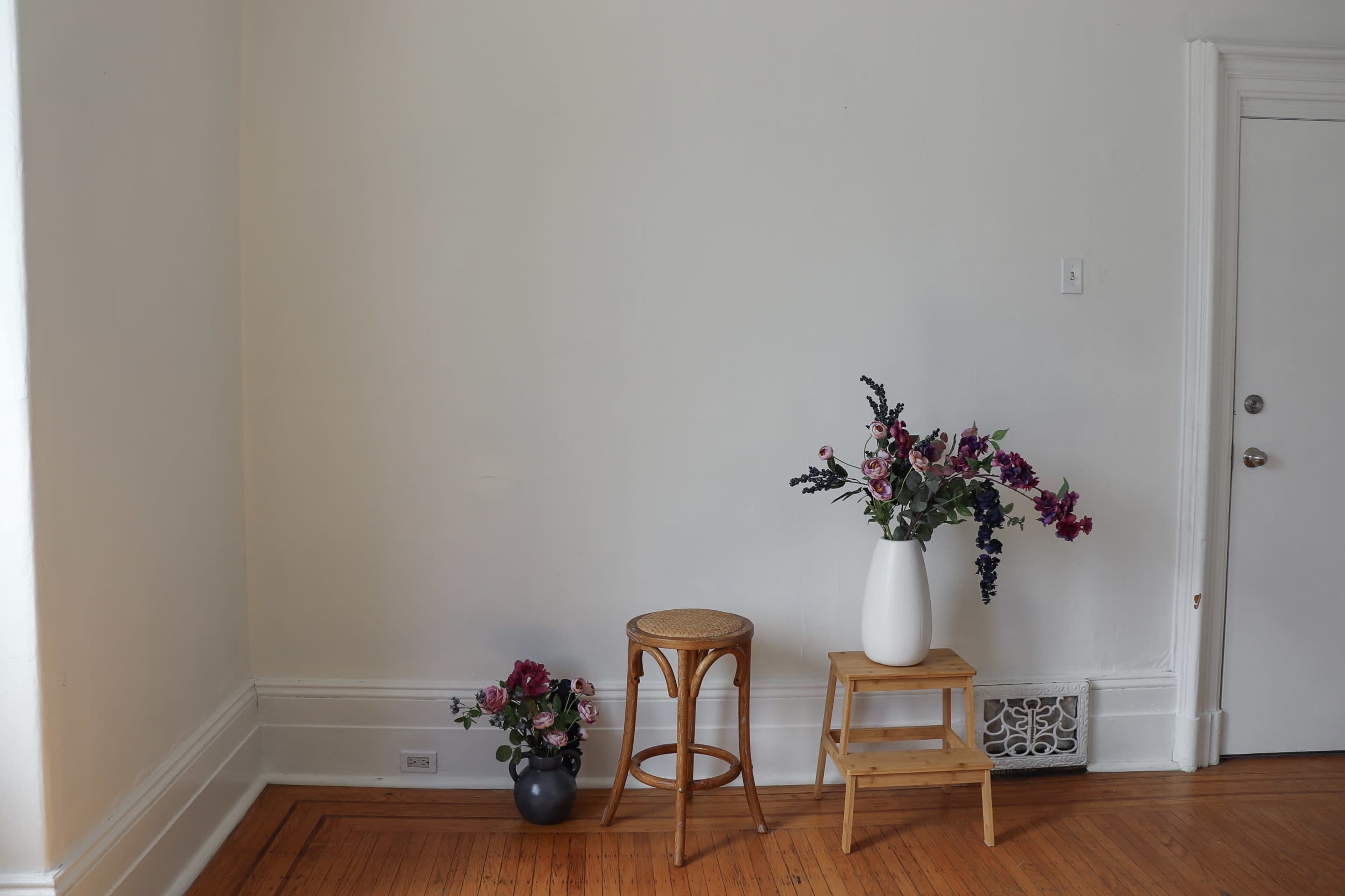A wooden stool and a small table with a tall vase of flowers are positioned against a bare white wall in a room with hardwood flooring.