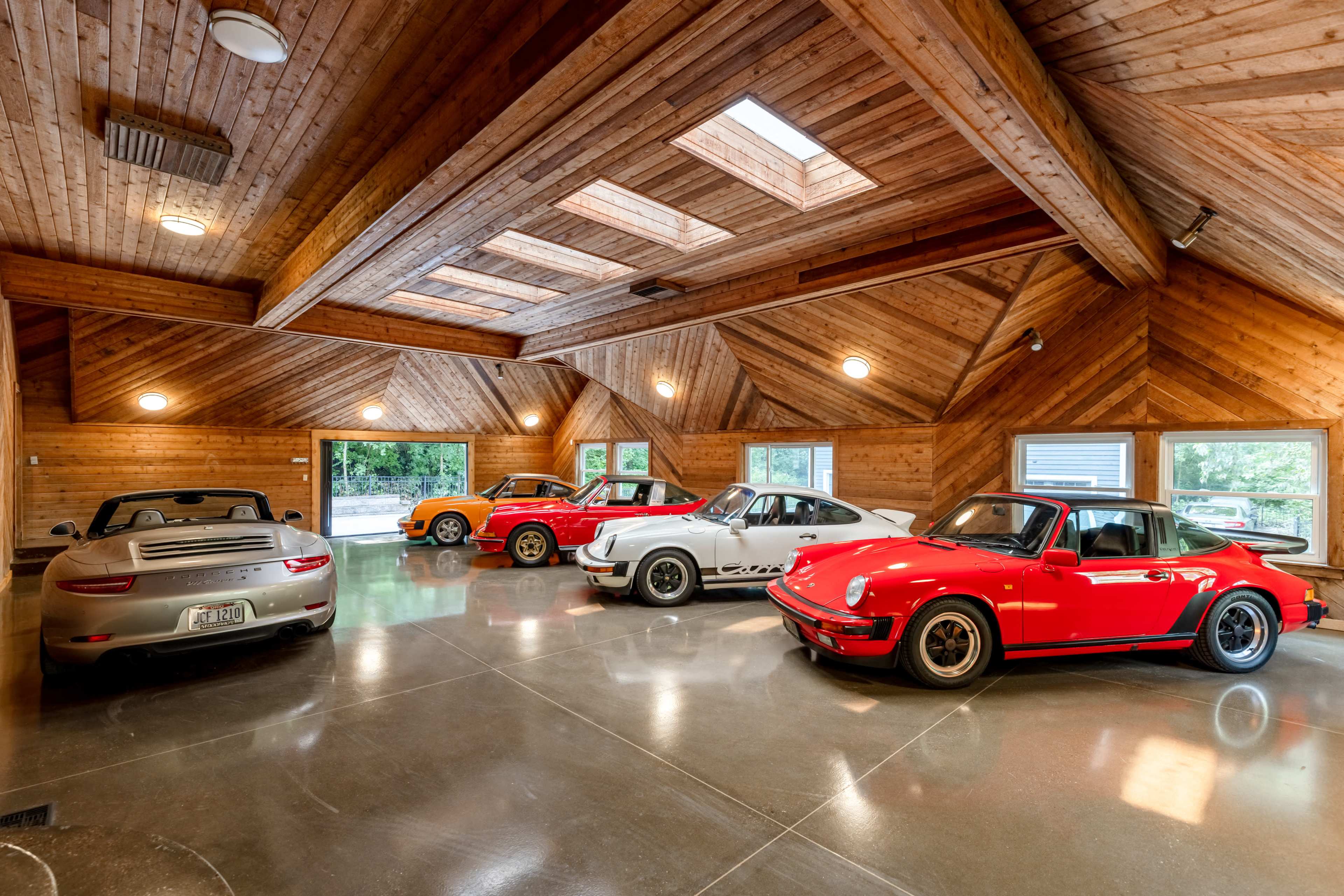 A spacious garage features four classic cars, including a red sports car and a silver convertible, illuminated by skylights and wooden paneling.