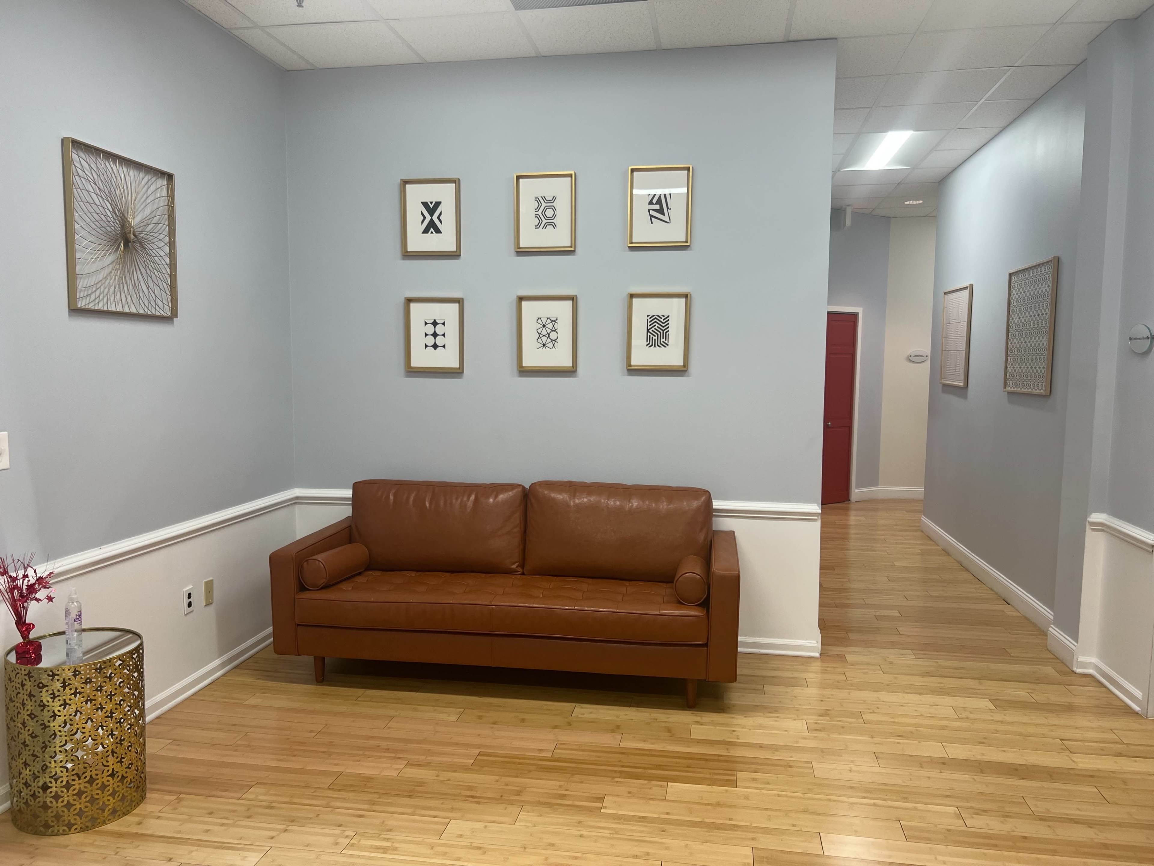 A brown leather couch is positioned against a light blue wall adorned with framed artwork, while a small decorative table with a floral arrangement sits nearby, leading into a well-lit hallway.