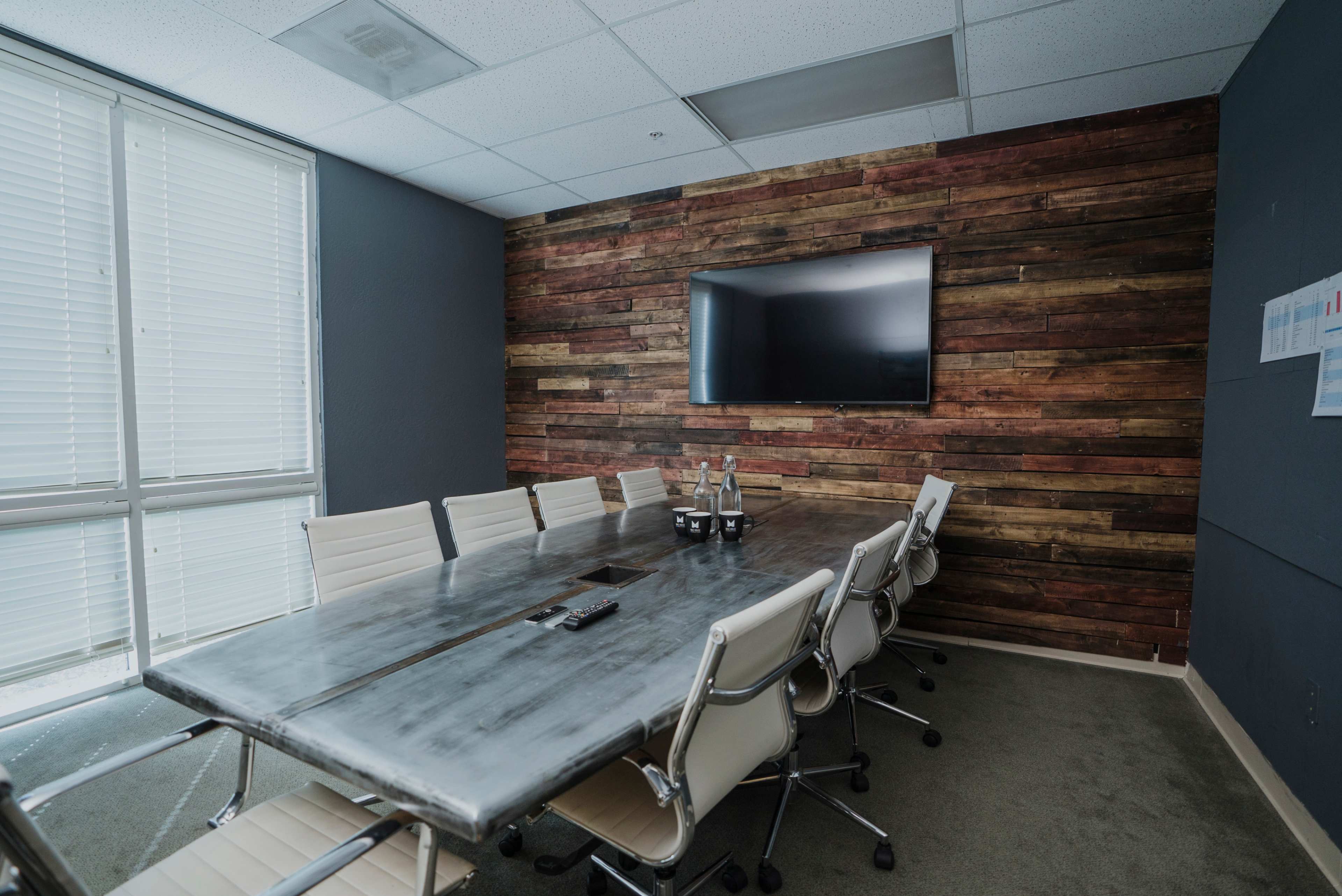 A conference room features a long metal table surrounded by white chairs, with a wooden accent wall and a wall-mounted television.