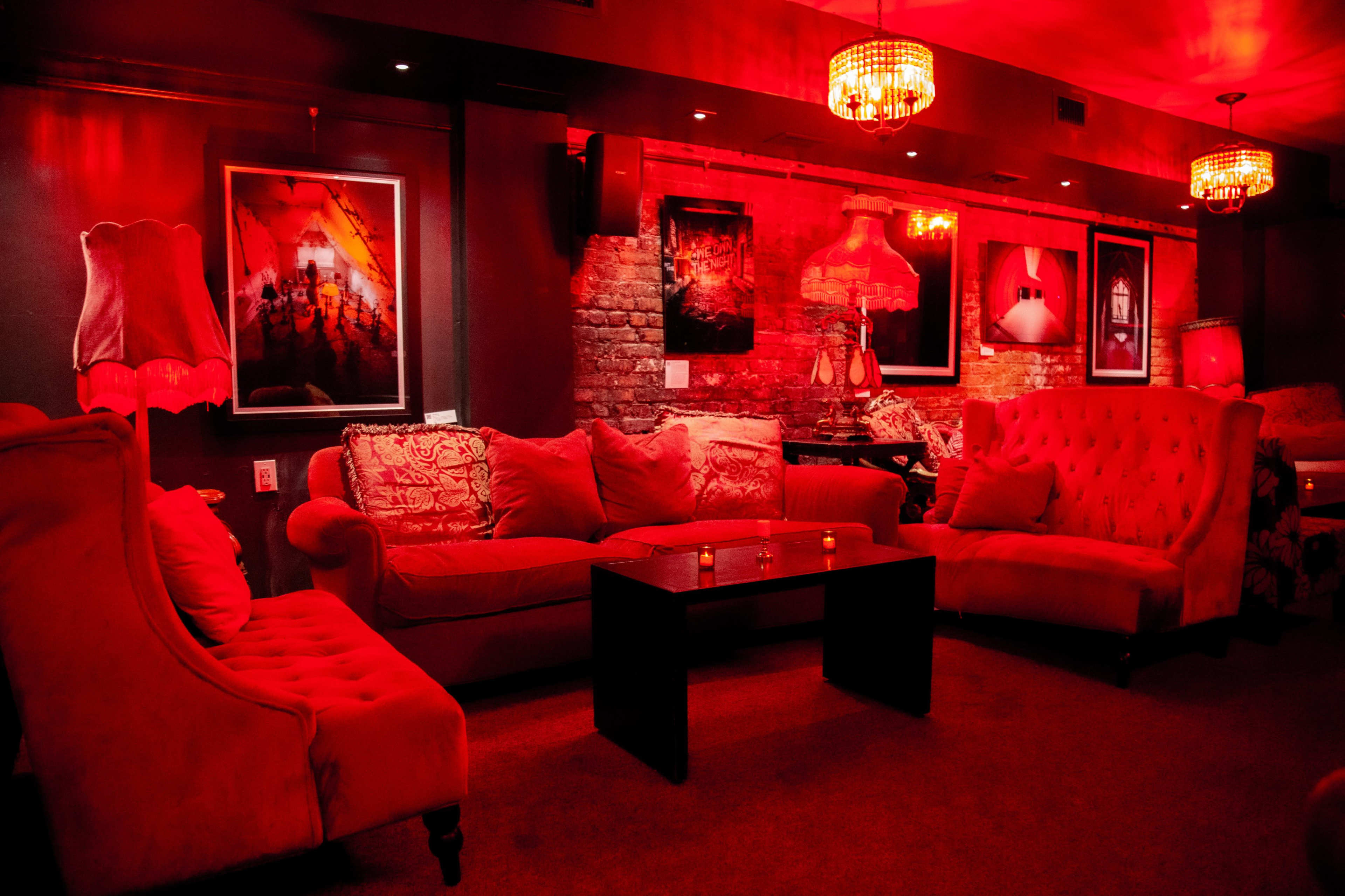 A dimly lit lounge area with red lighting, featuring plush red sofas, a dark coffee table, and framed artwork on the walls.